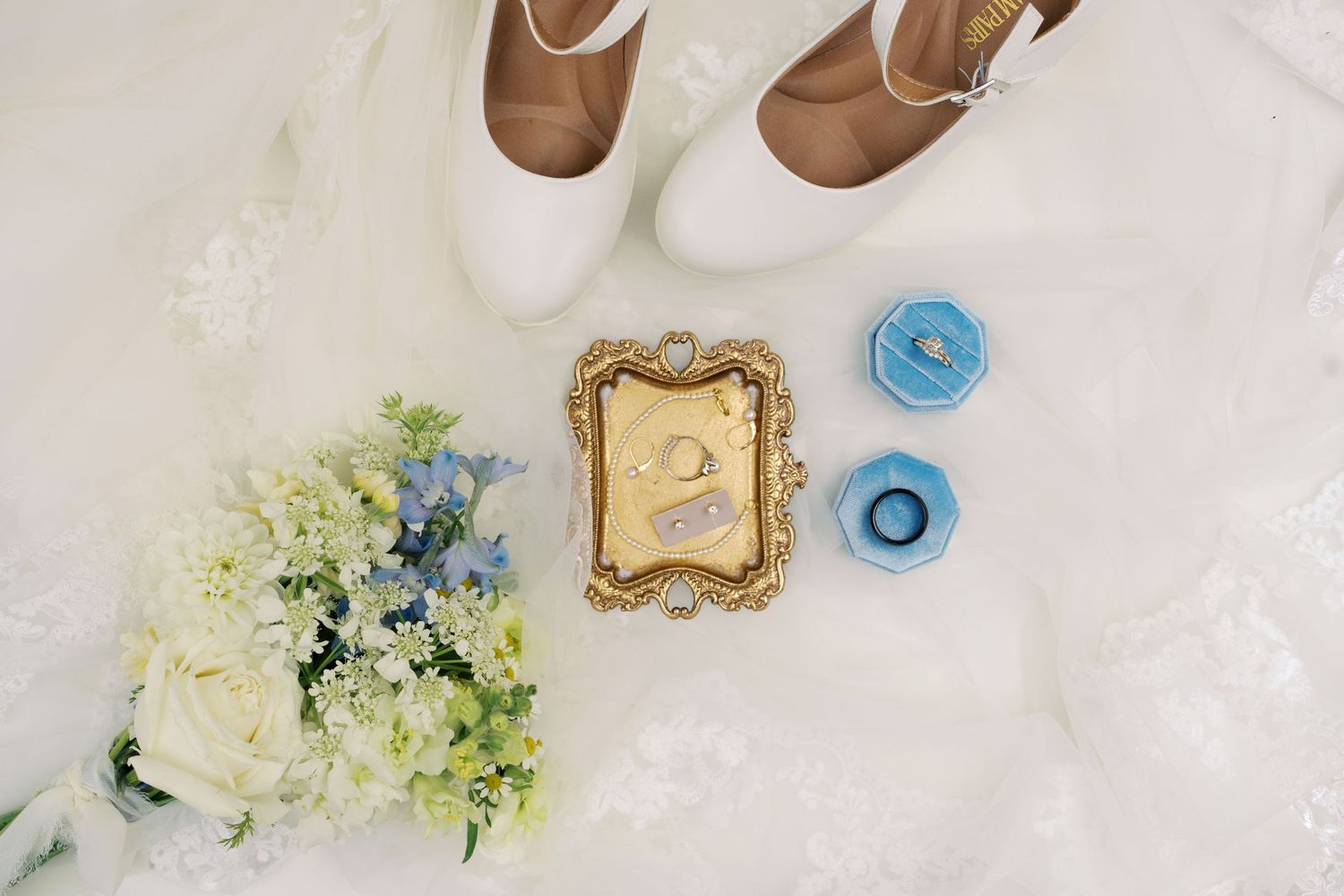 Bridal flat lay with white heels, gold ring dish, blue velvet ring boxes, and a white and blue floral bouquet on lace.