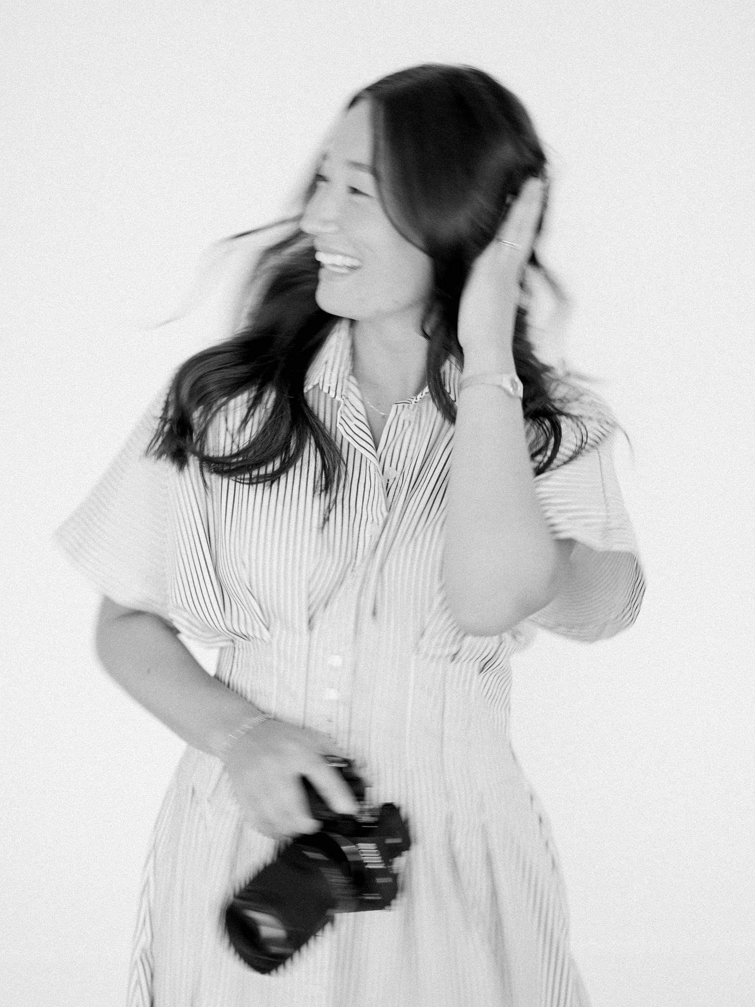Black and white photo of a smiling woman in a striped dress holding a camera, hair blowing in the wind.