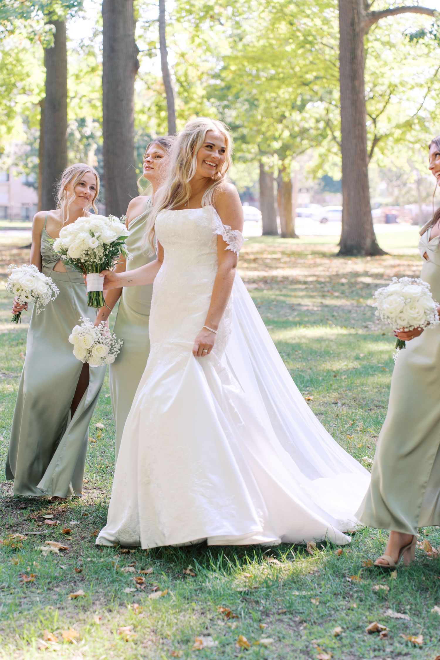 Bridesmaids in sage green dresses pose playfully with the bride in a white gown outdoors among trees.