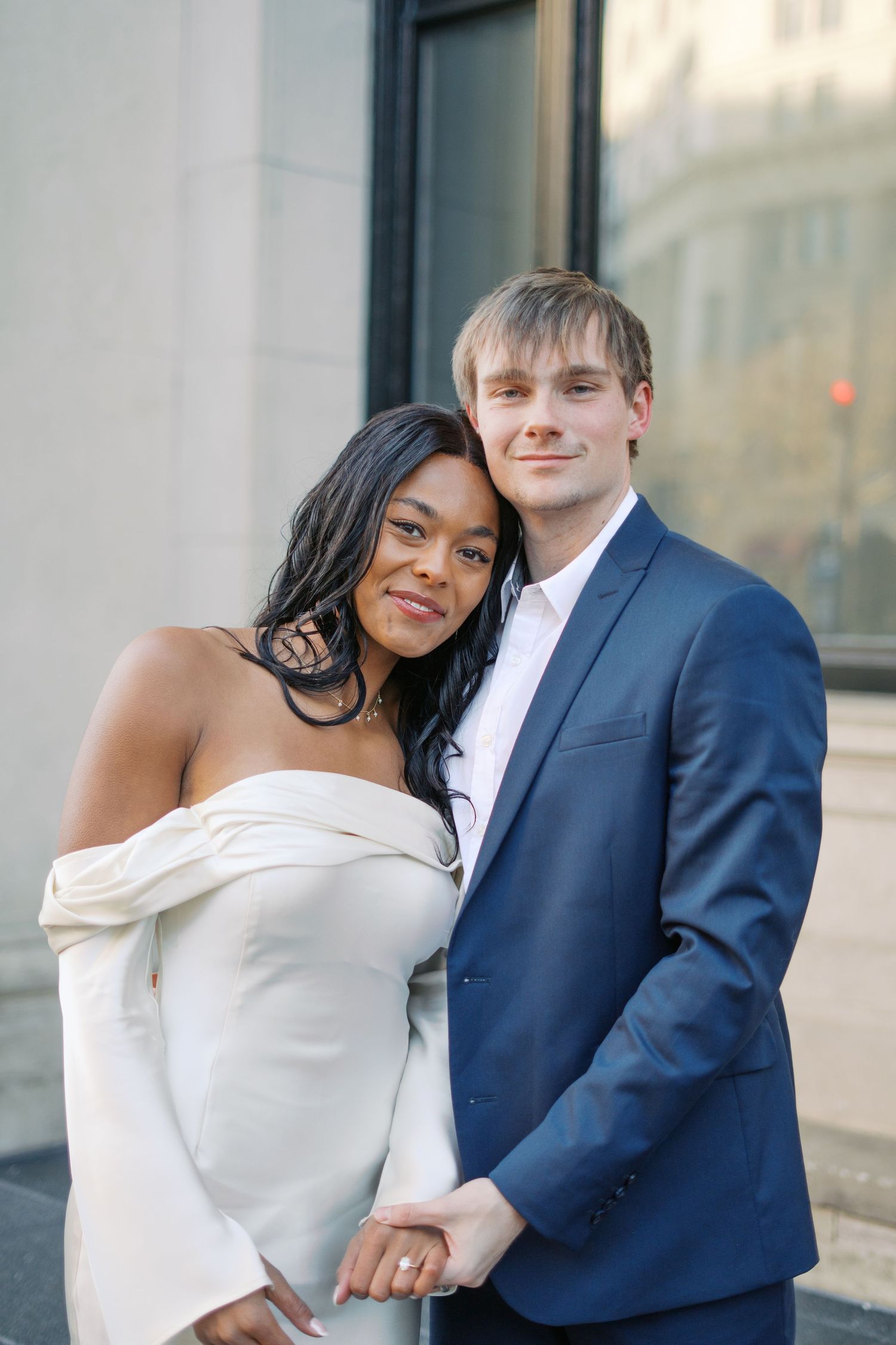 A couple in formal attire embraces and smiles against a white building exterior.