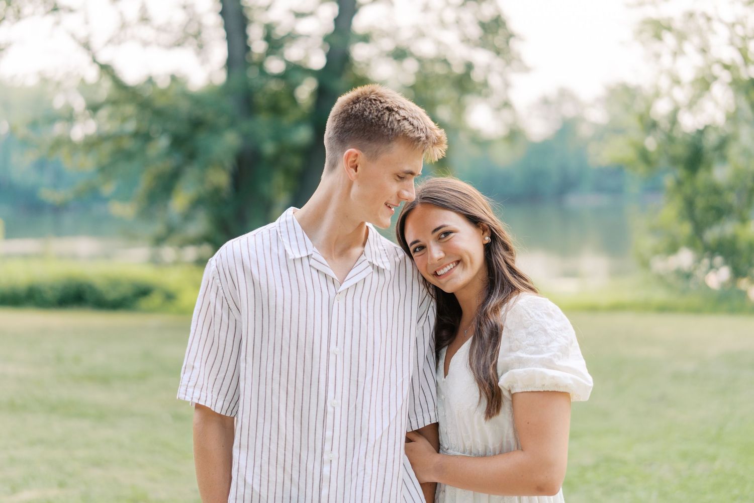 A happy young couple smiles together outdoors near a lake, with lush green trees in the background.