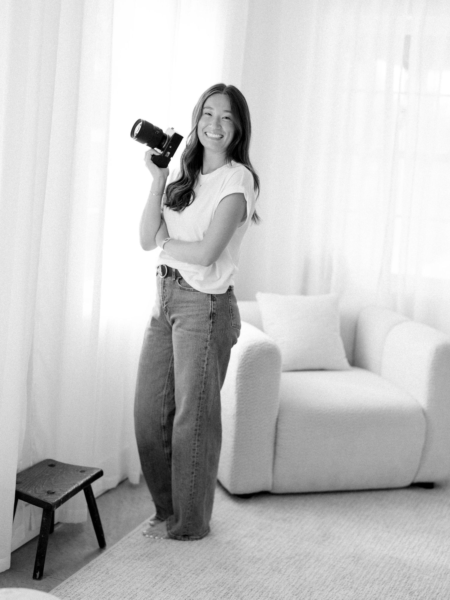 Black and white photo of a smiling woman holding a camera, standing in a bright minimalist room with sheer curtains.