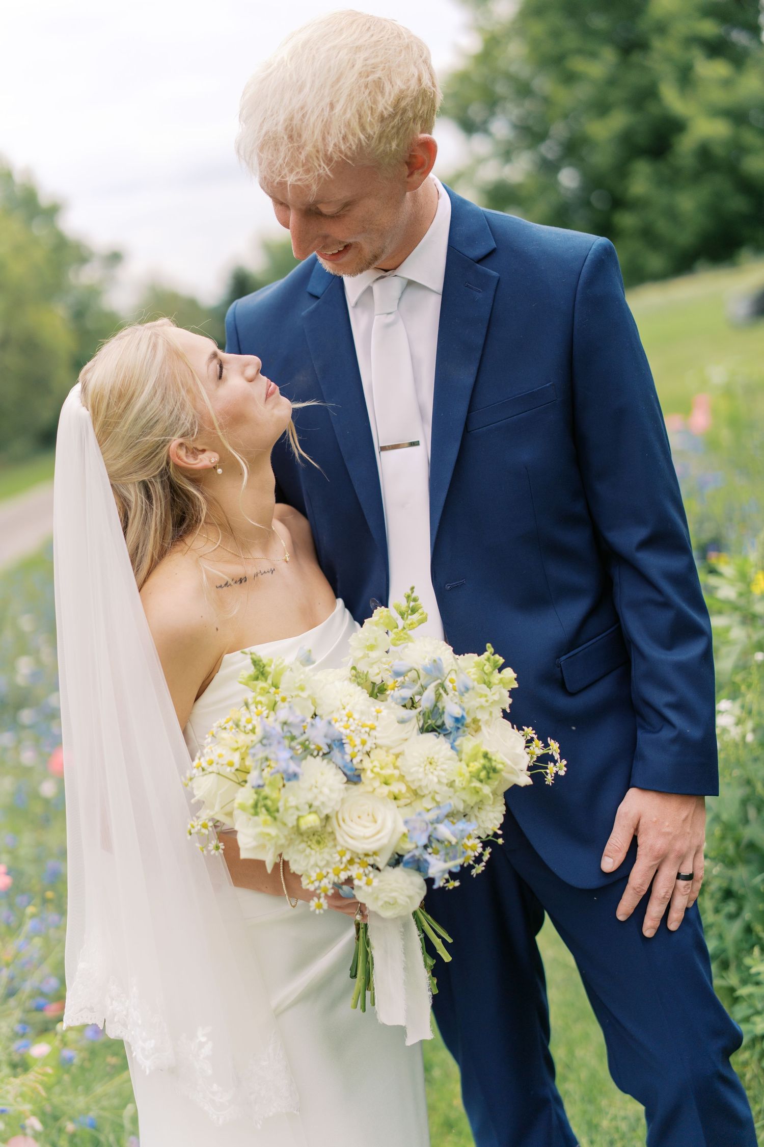 Bride and groom share a romantic moment outdoors, she holds a blue and white bouquet, he wears a navy suit.