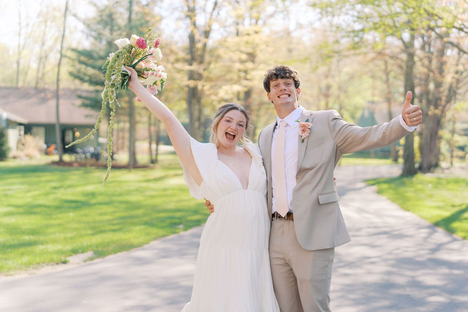 Joyful newlyweds celebrate outdoors, bride raising bouquet and groom giving thumbs up in spring garden setting.