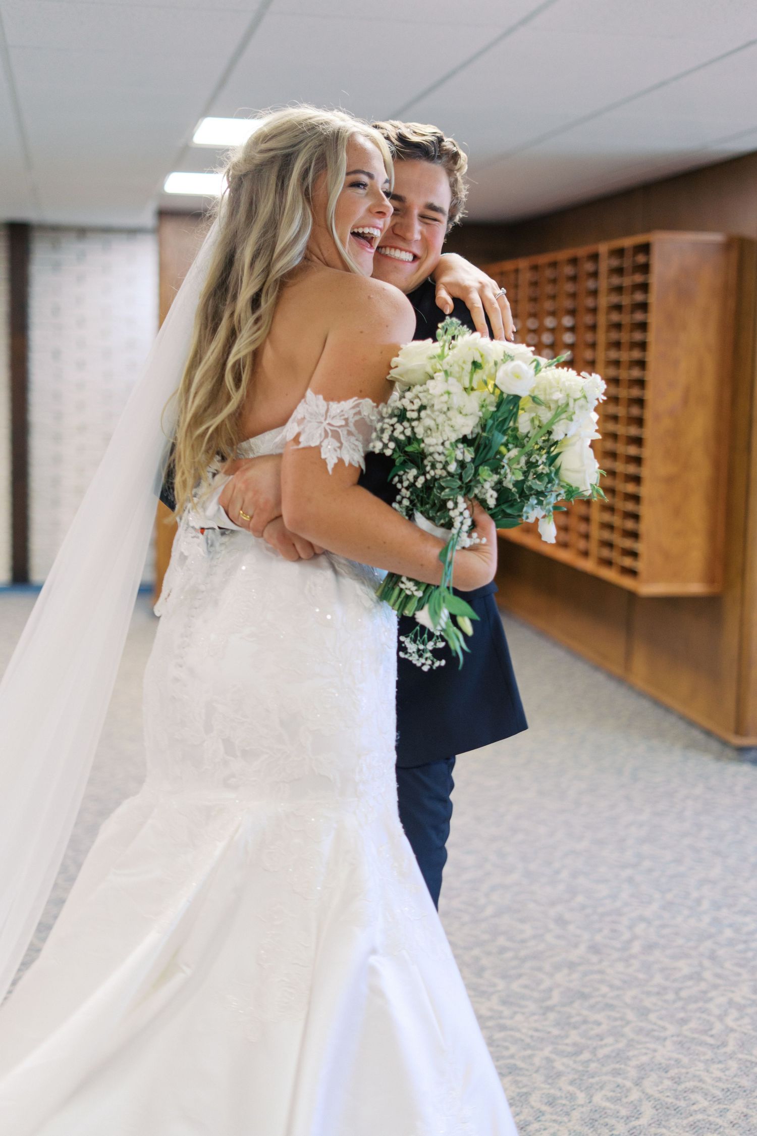 A joyful wedding couple embraces in a church hallway while holding a white bouquet.