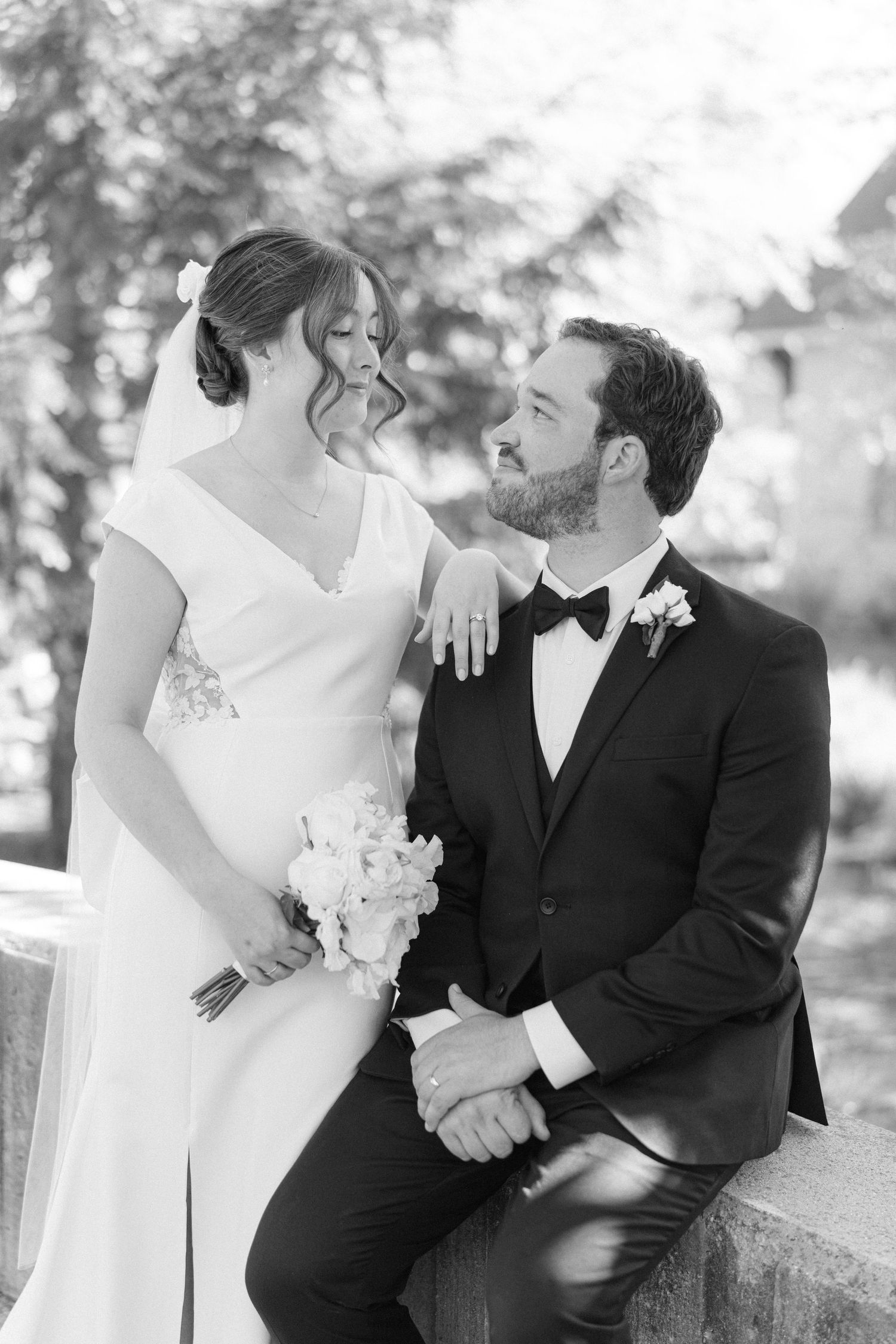 Black and white wedding photo of bride holding bouquet standing beside seated groom in tuxedo outdoors.
