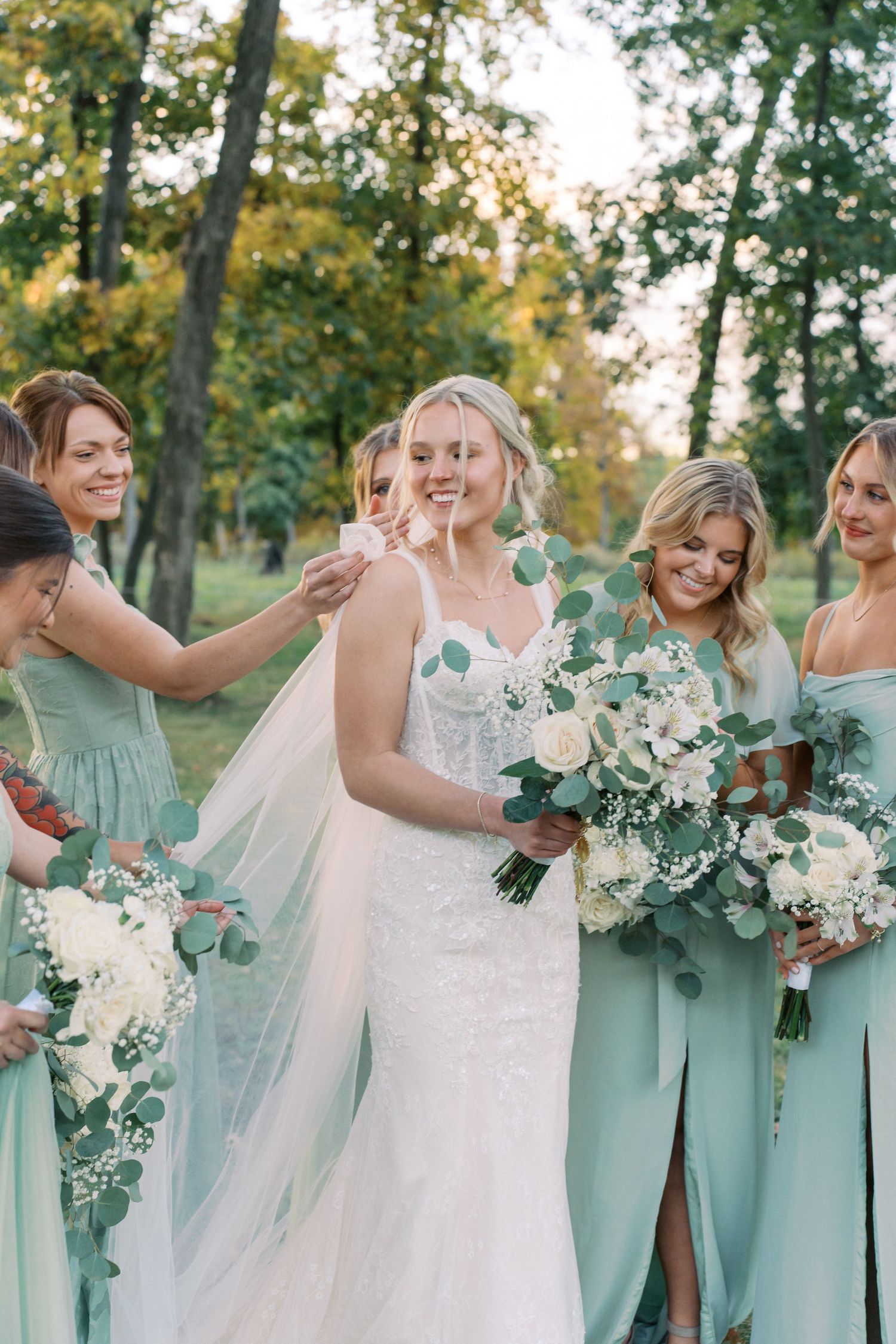 Bridesmaids in mint green dresses gather around bride holding white floral bouquets in outdoor setting.