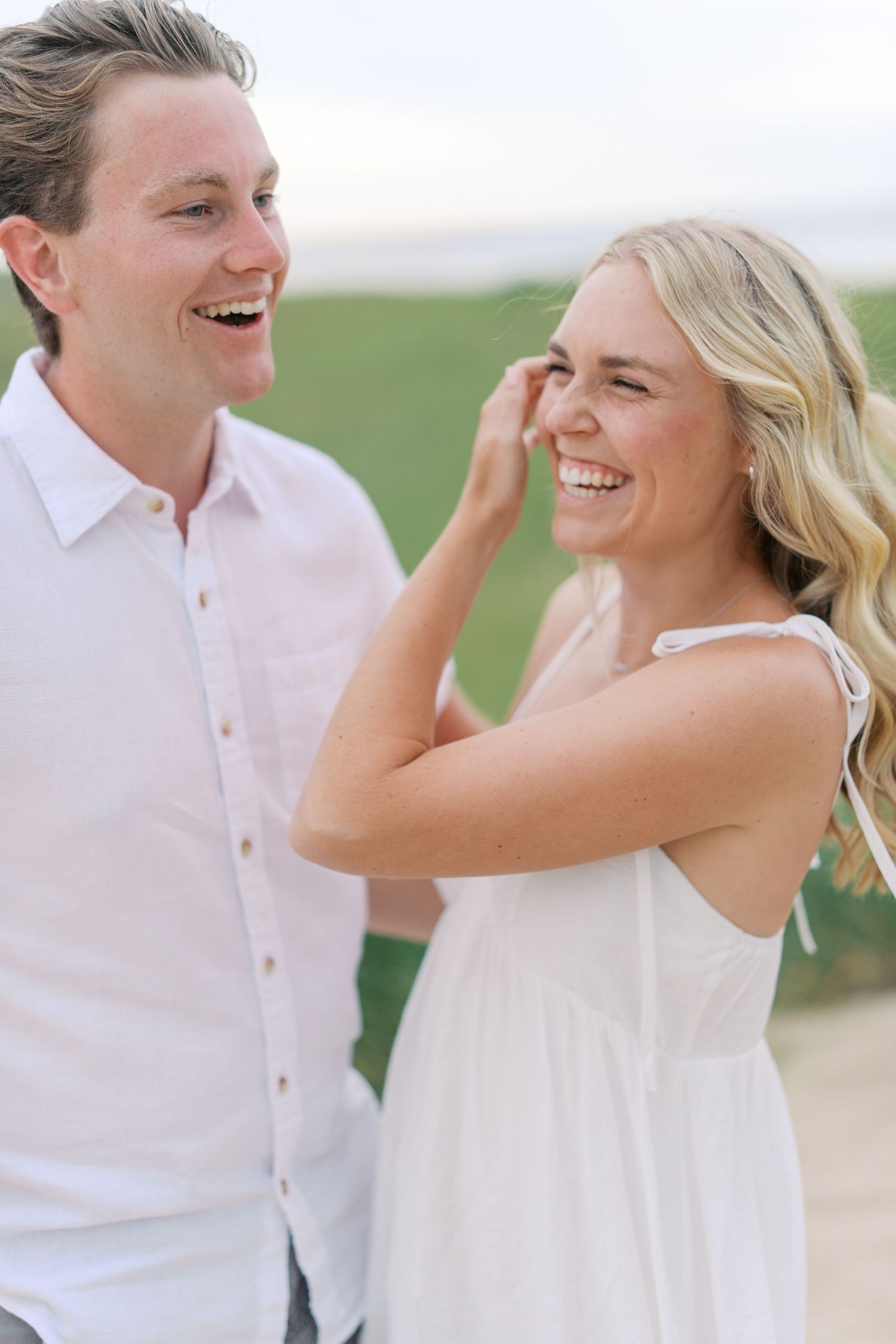 A laughing couple in white outfits share a joyful moment outdoors, with the woman tucking her blonde hair behind her ear.