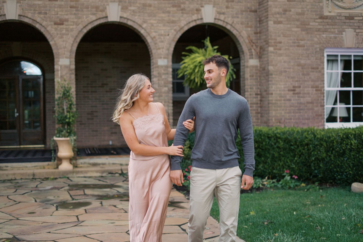 A couple stands together on stone steps with brick arches and landscaping.