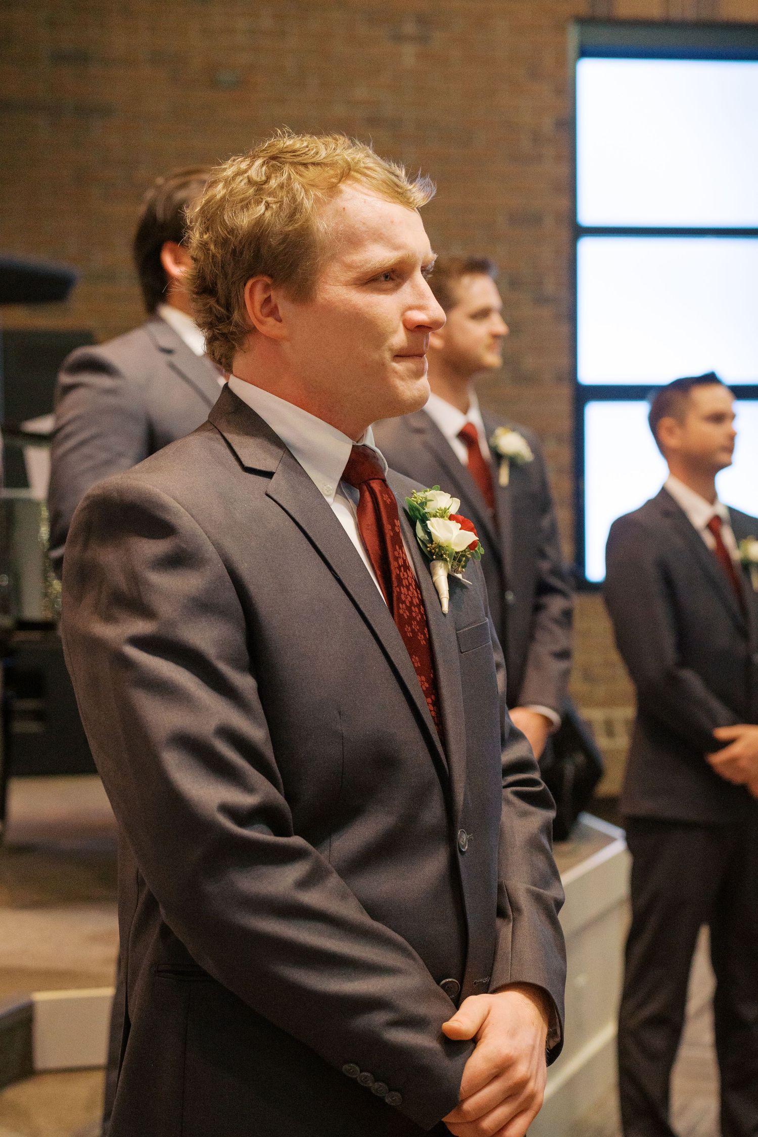 Groomsmen in dark suits with burgundy ties stand during wedding ceremony.