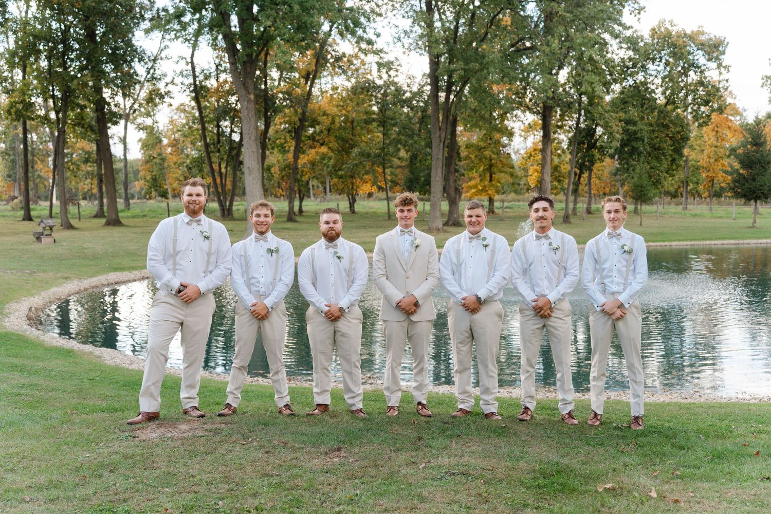 Wedding party in white and mint green attire poses by a lakeside on a sunny autumn day.