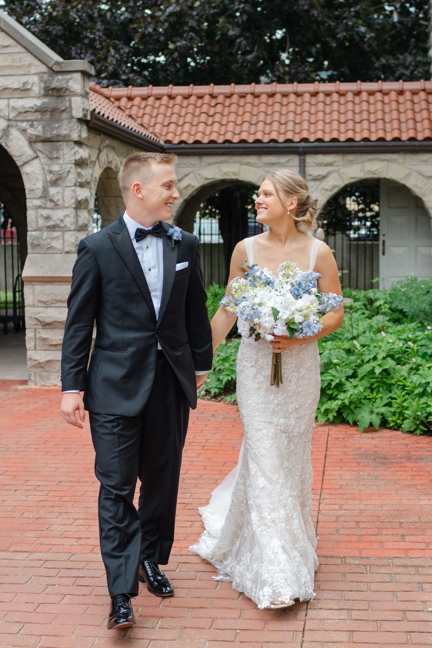 Wedding couple walking and posing along a brick pathway near classical architecture and gardens.