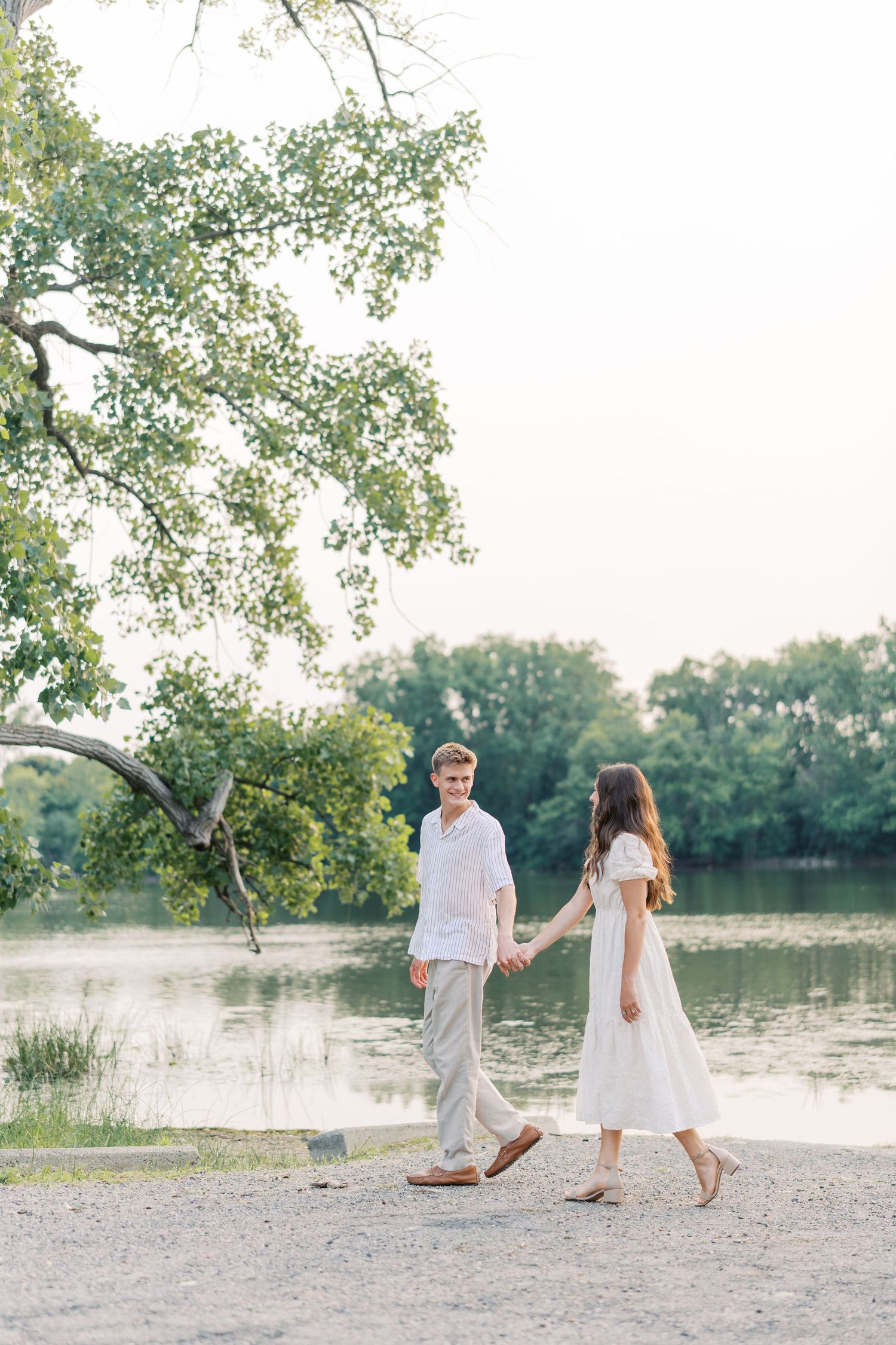 Couple in white outfits holding hands and walking by a serene lake surrounded by lush green trees.