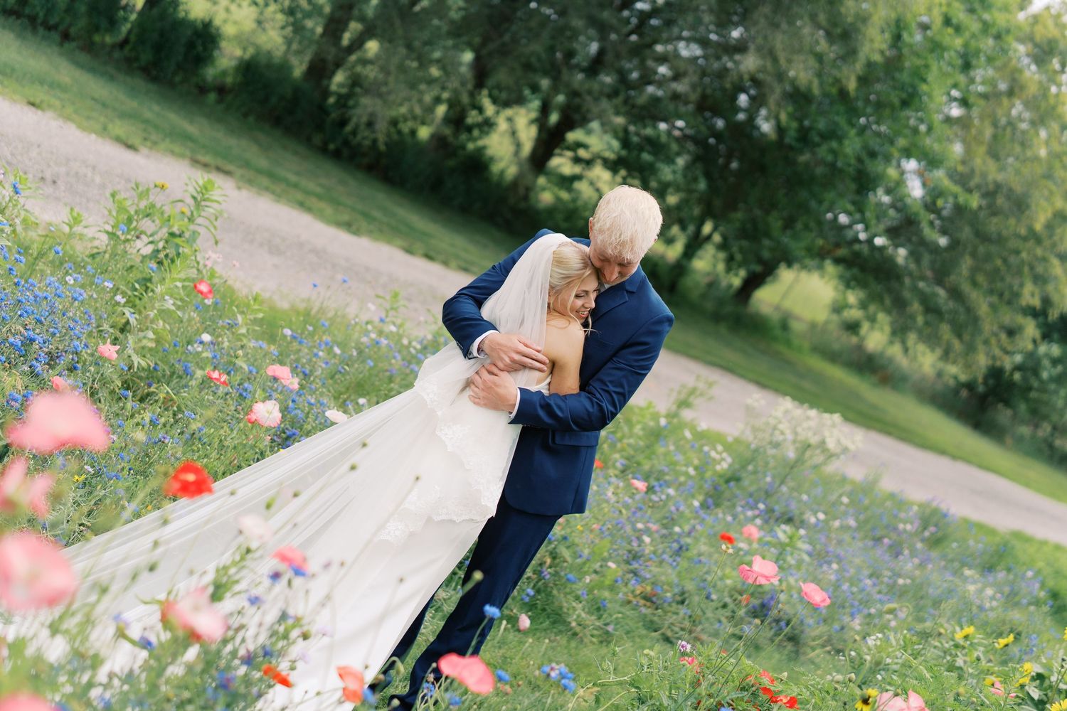 Bride and groom embracing in a colorful wildflower meadow, groom in navy suit, bride in white gown with veil.
