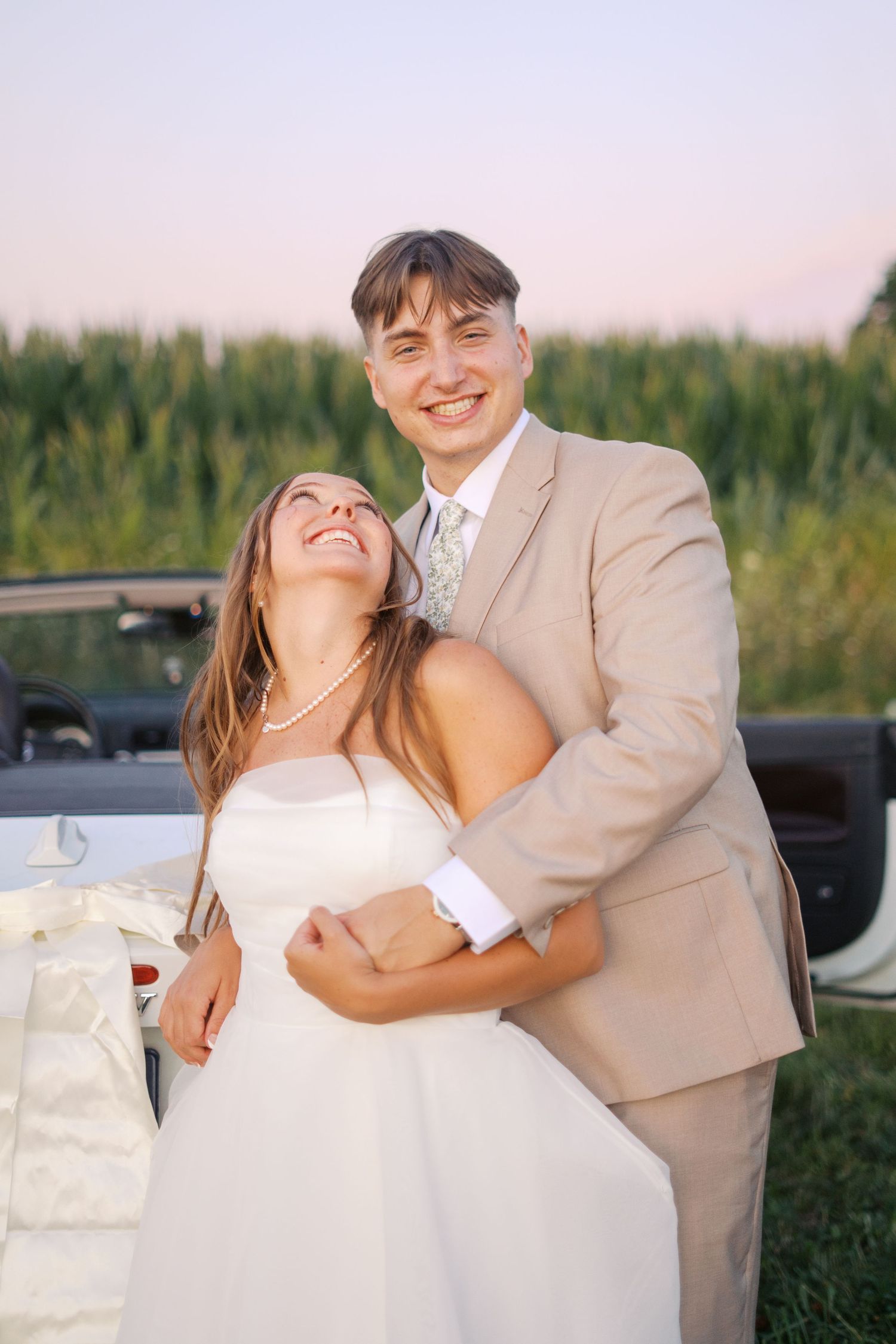 A joyful wedding couple shares laughter while posing by their car outdoors.
