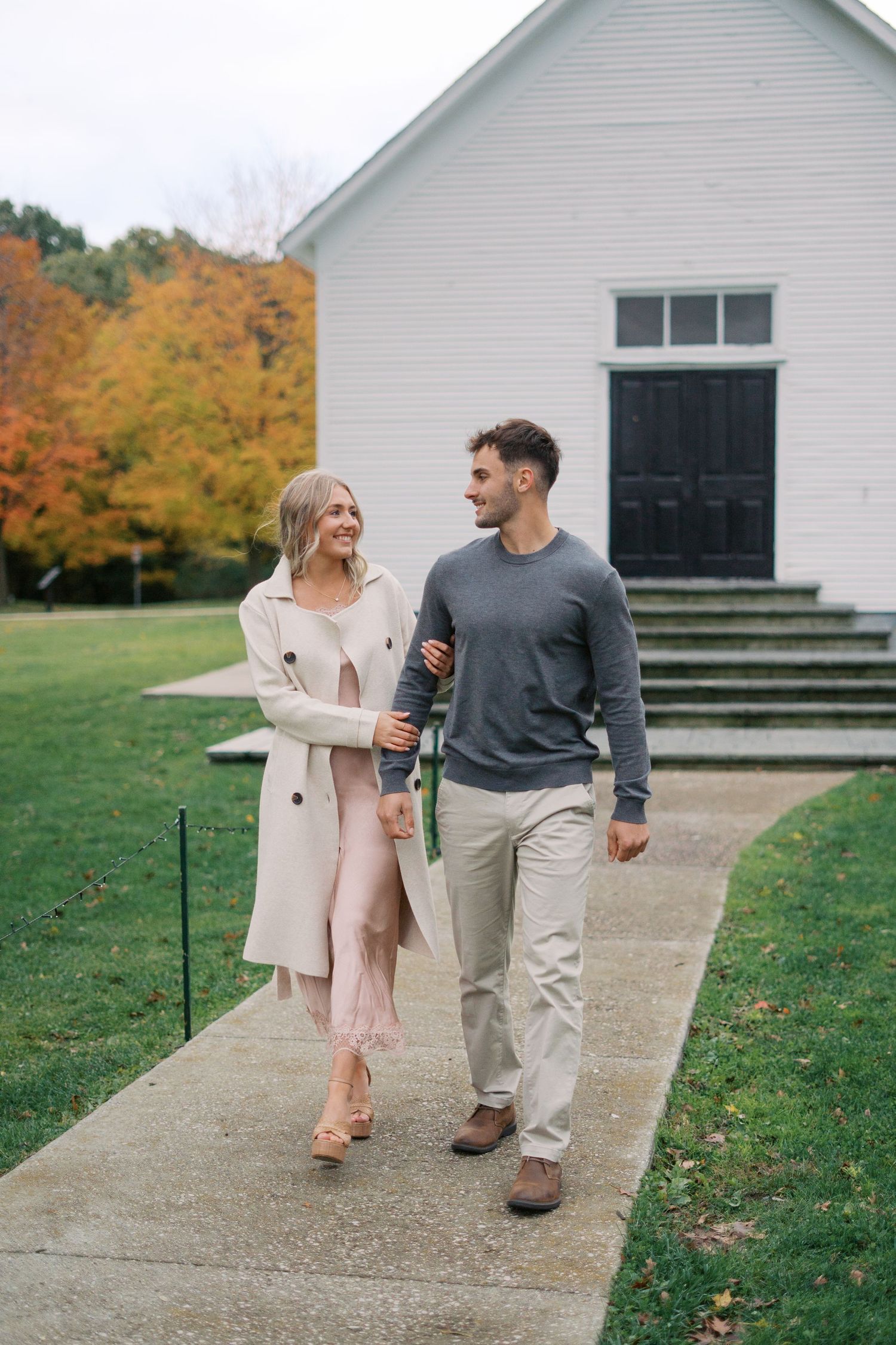 A couple walking together on a path near a white church building during fall.