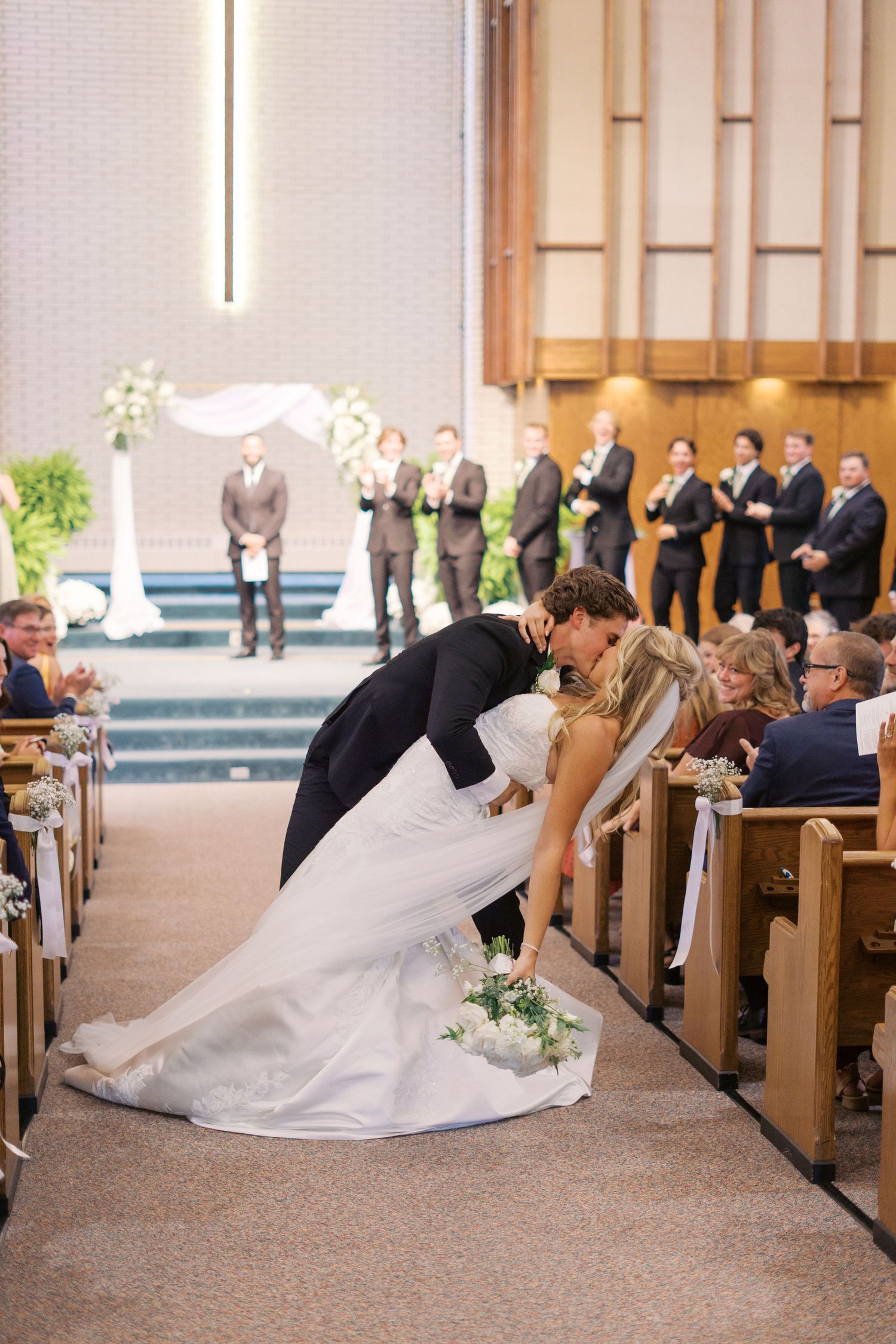 A wedding couple shares a romantic dip pose in a church aisle surrounded by guests.