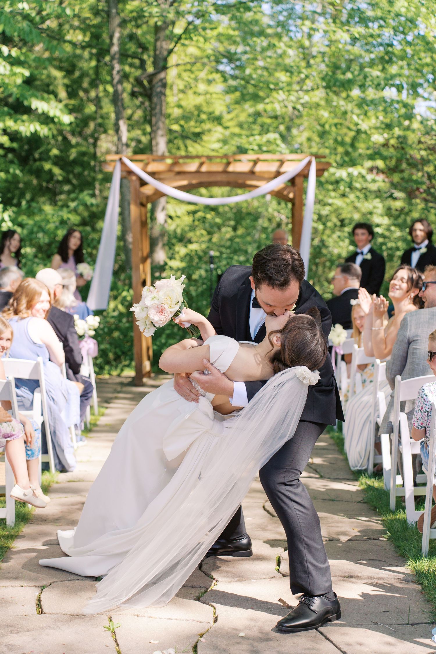 Groom dips bride for a kiss at outdoor wedding ceremony with white chairs and wooden arch.
