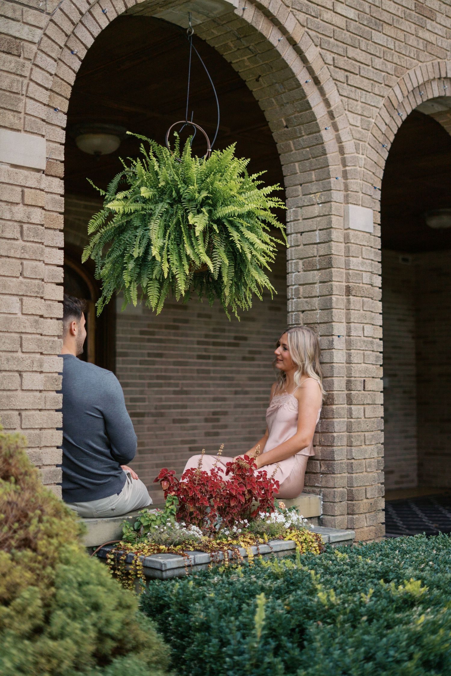 Two people sit together under decorative brick archways with hanging ferns.
