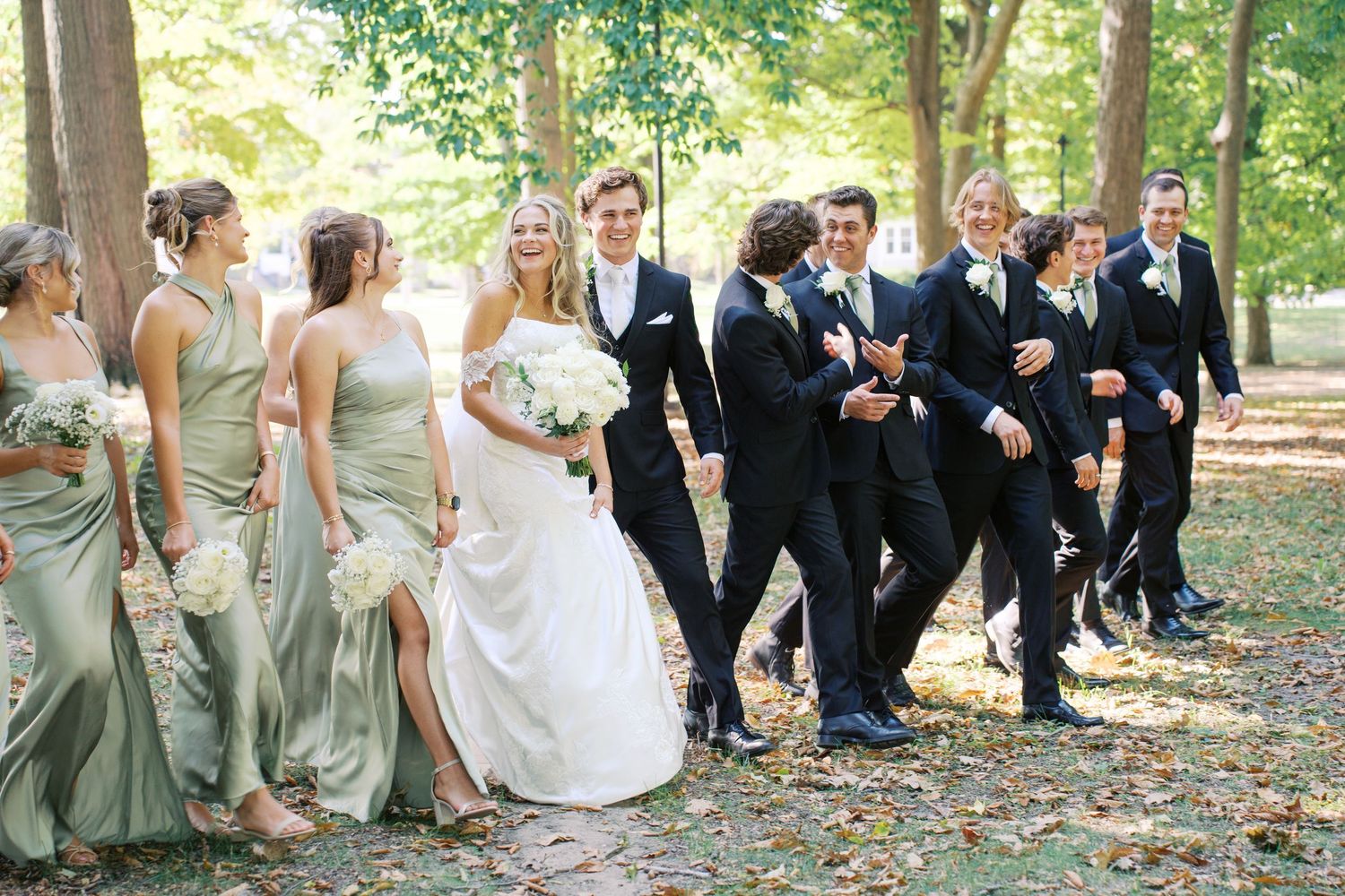 Wedding party in sage green dresses and black tuxedos poses casually on grass under trees.