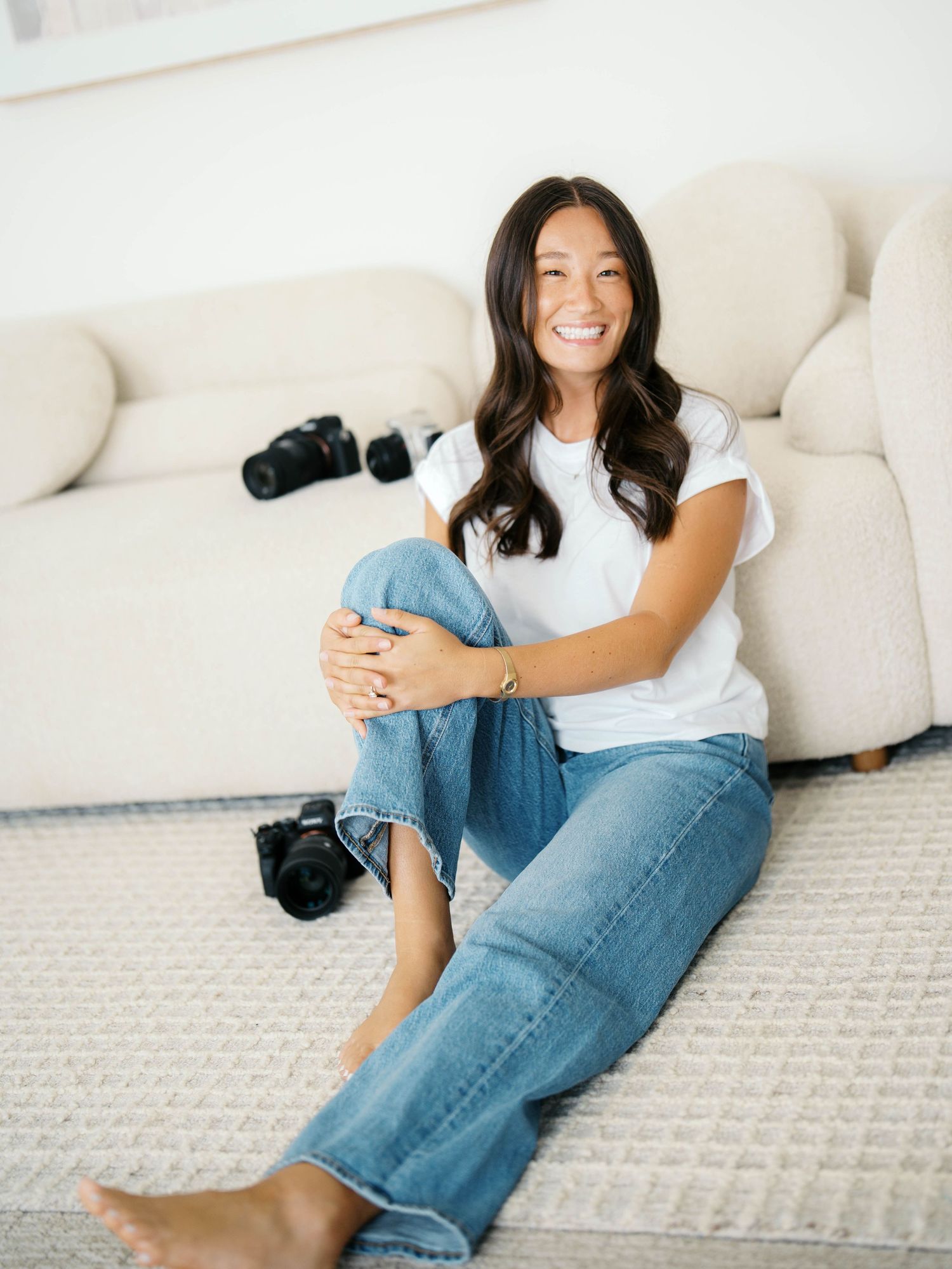 Smiling woman in white tee and jeans sits on floor near sofa with cameras, bright airy room.
