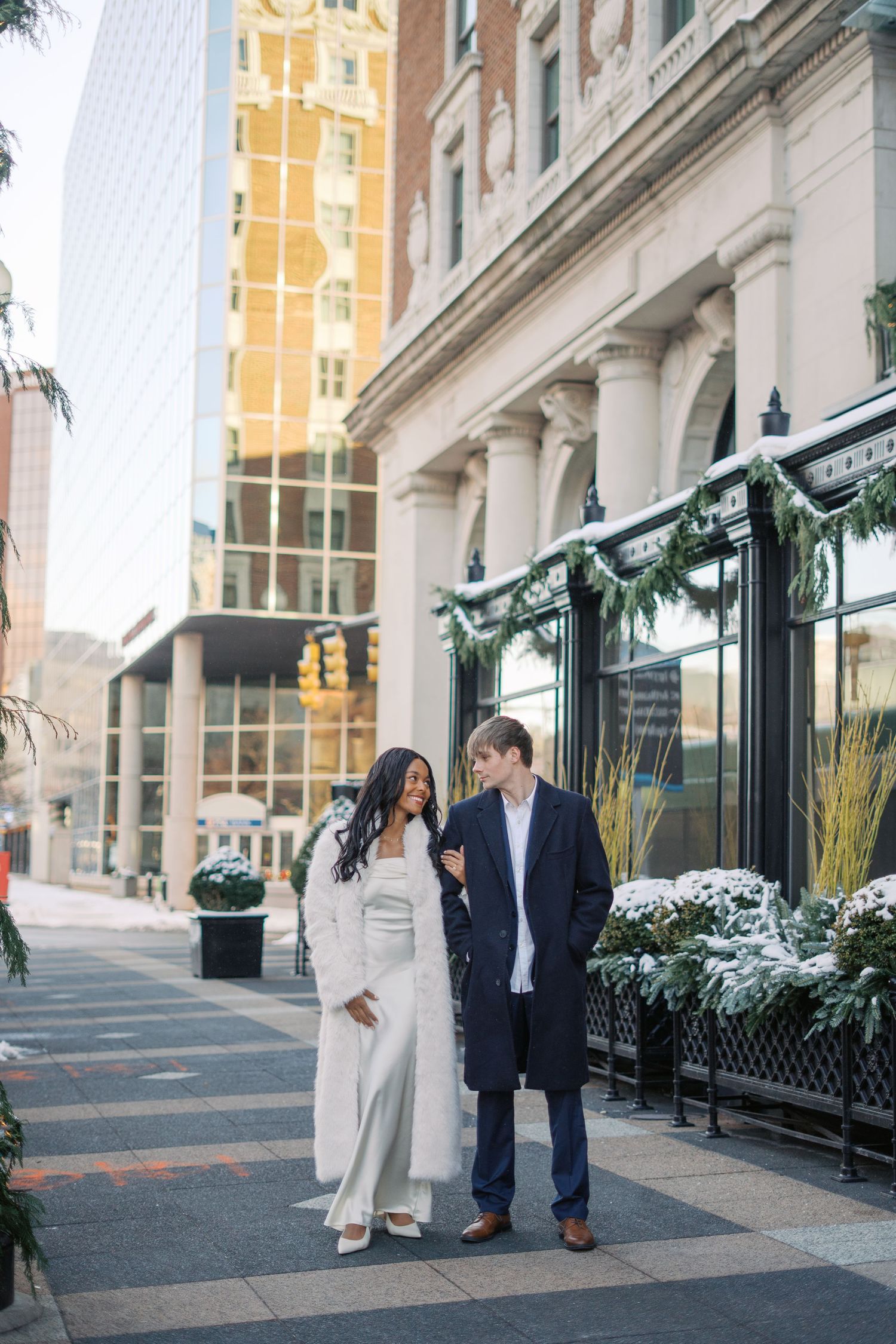 A couple poses outside a modern building with snowy winter decorations.