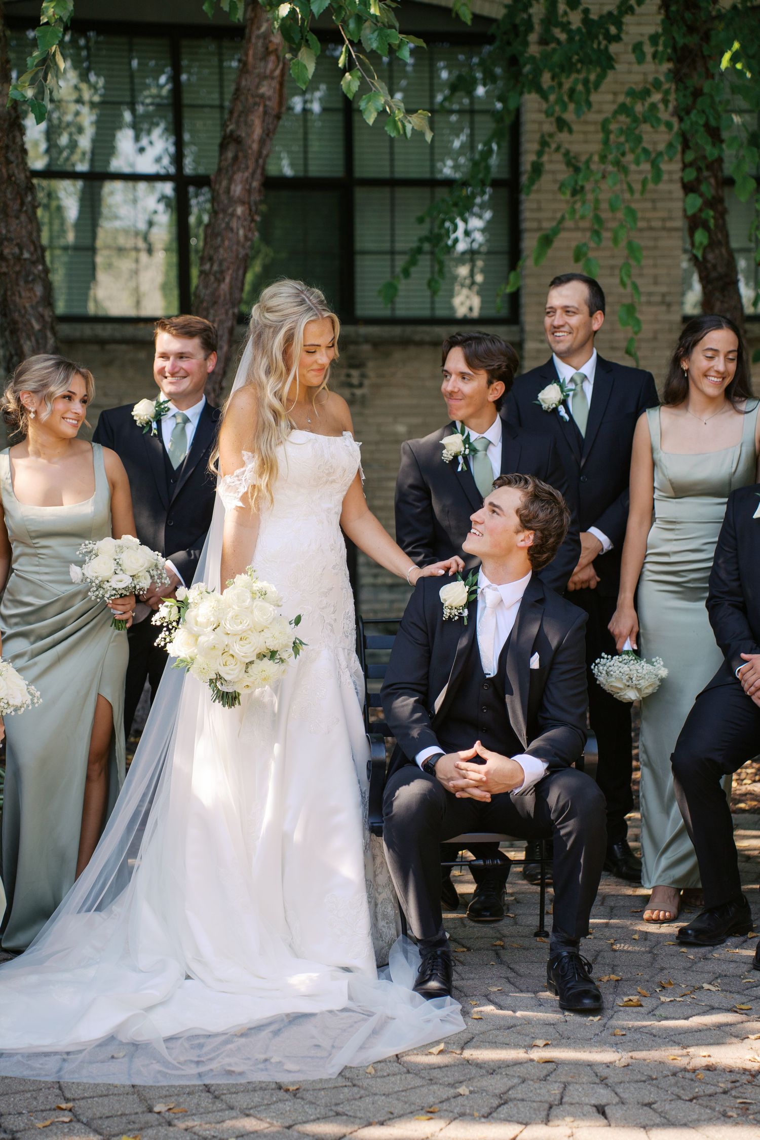 Wedding party gathered outdoors with bridesmaids in gray dresses and groomsmen in dark suits on brick patio.