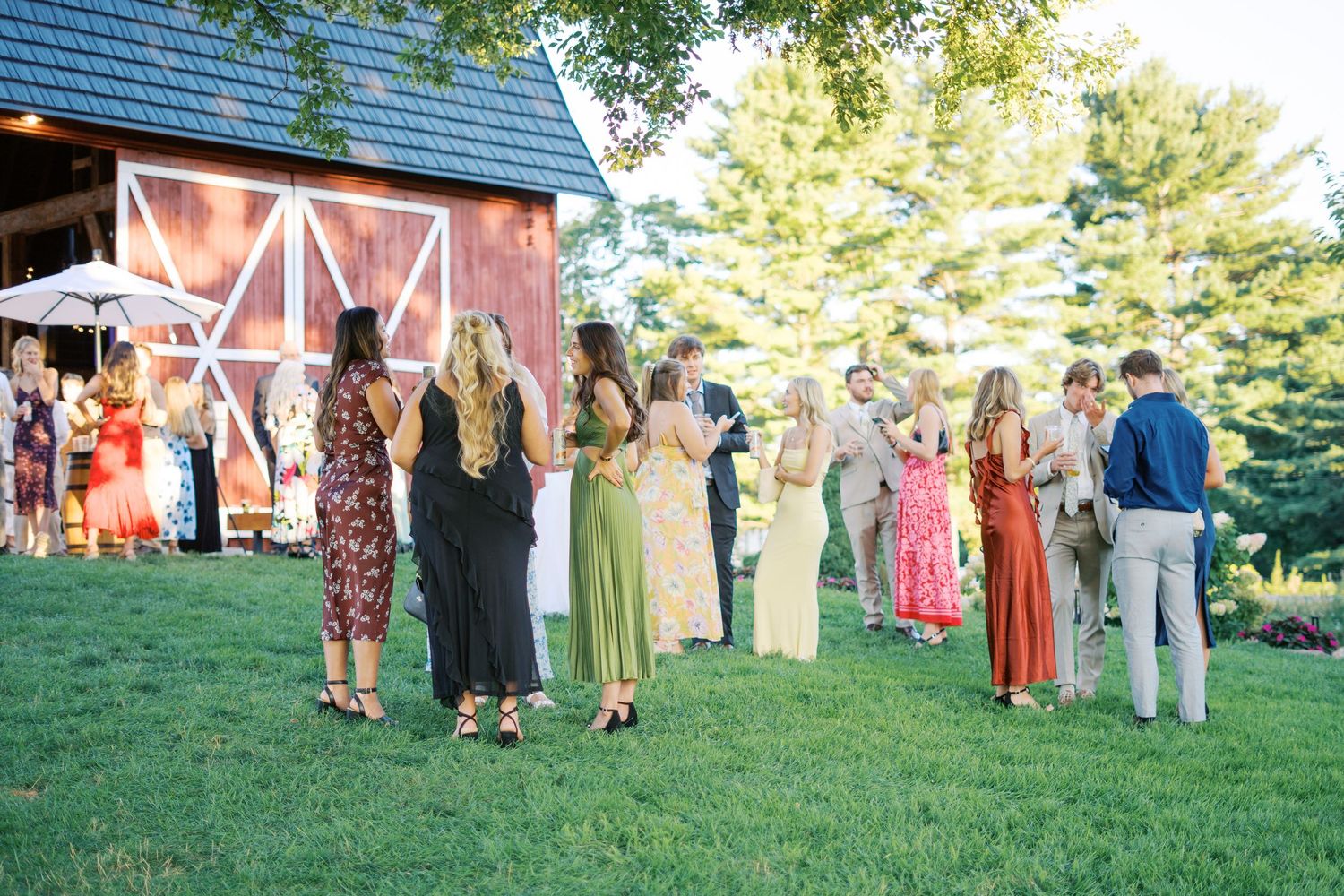 Wedding guests in colorful dresses gather outside a red barn on a sunny day.