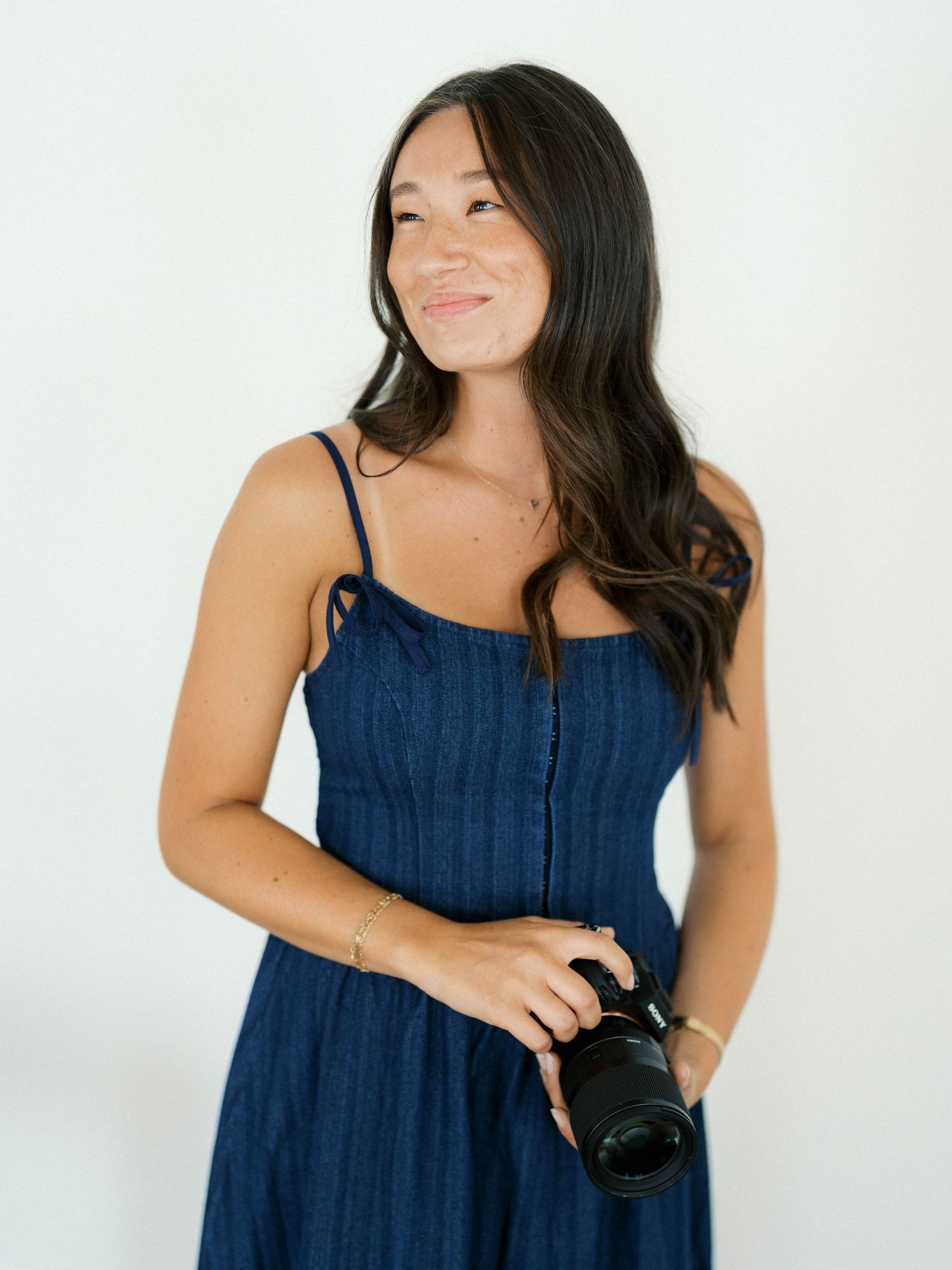 A smiling young woman in a navy blue sundress holds a DSLR camera against a white background.