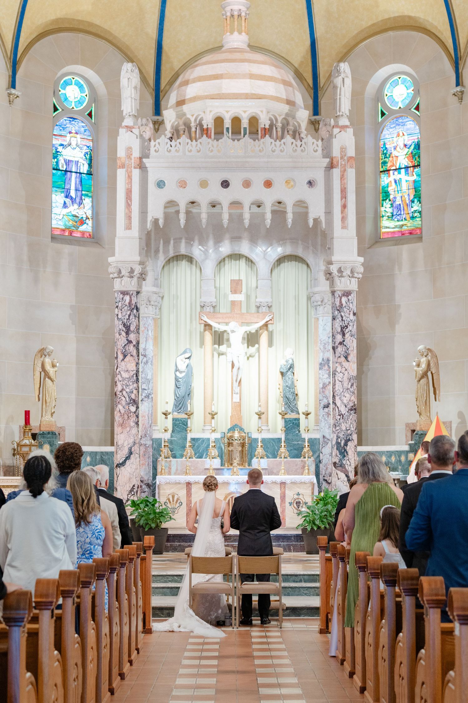 A wedding ceremony taking place in a beautiful Catholic church with stained glass windows and ornate marble altar.