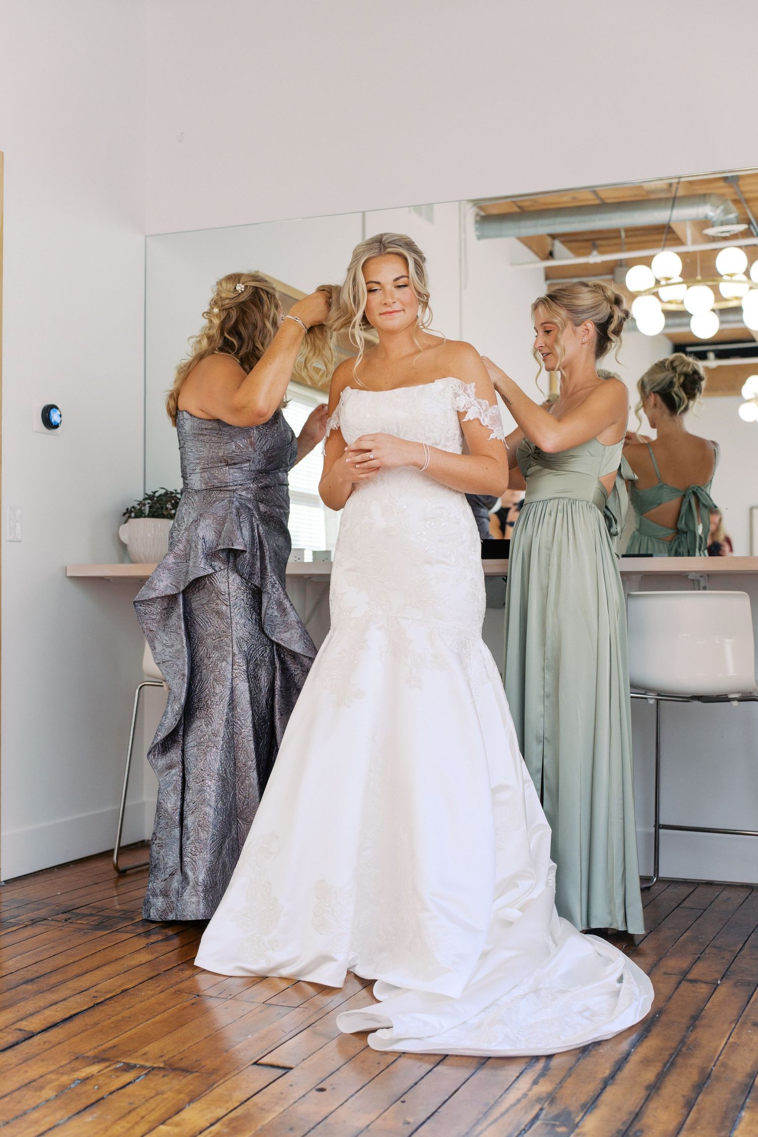 Wedding party helping with dress preparations in a bright room with wooden floors.