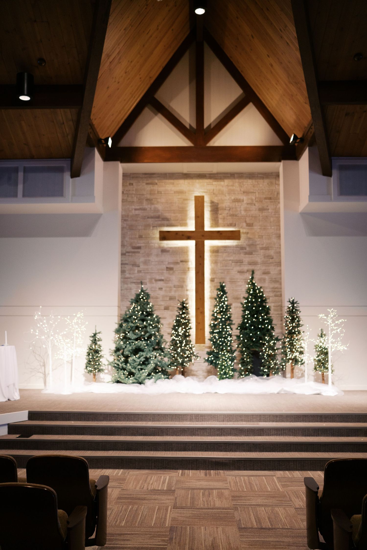 Modern church altar decorated with illuminated Christmas trees and a backlit cross on stone wall.