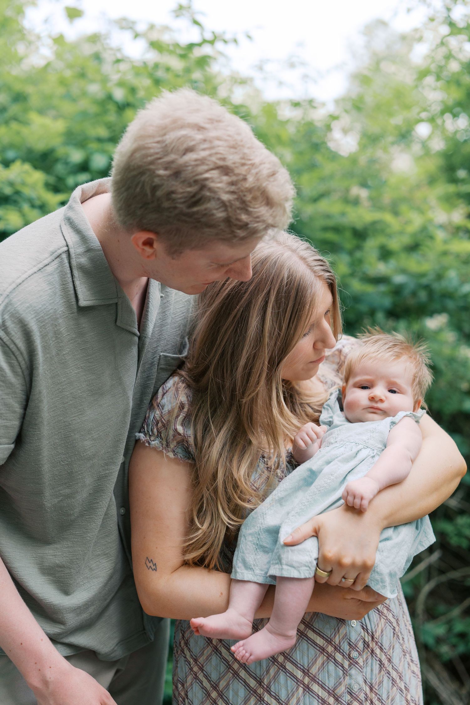 A couple shares a tender moment kissing over their newborn baby outdoors with lush green foliage behind them.