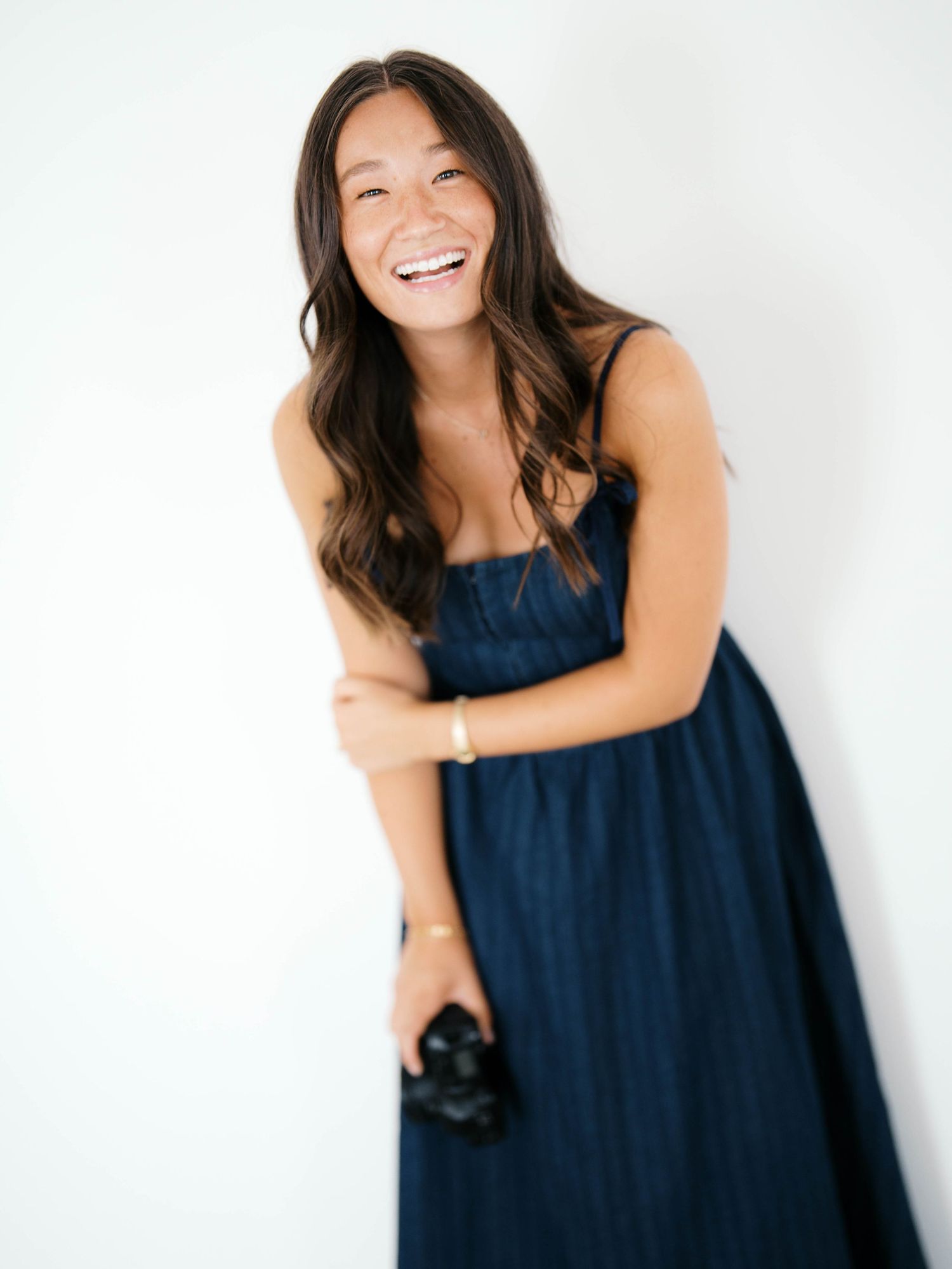 Smiling woman in navy blue sundress holding a camera, posing against a white background with long wavy hair.