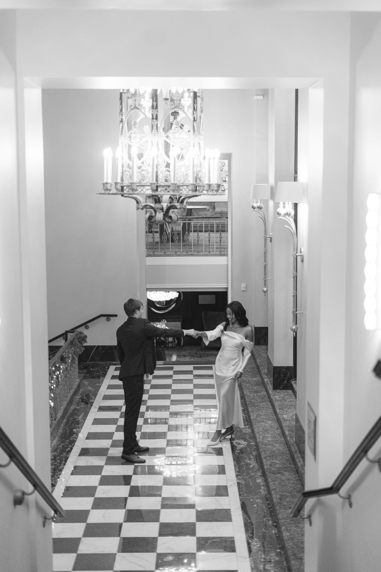 A couple dances dramatically on a checkerboard floor in an elegant hallway with chandeliers.