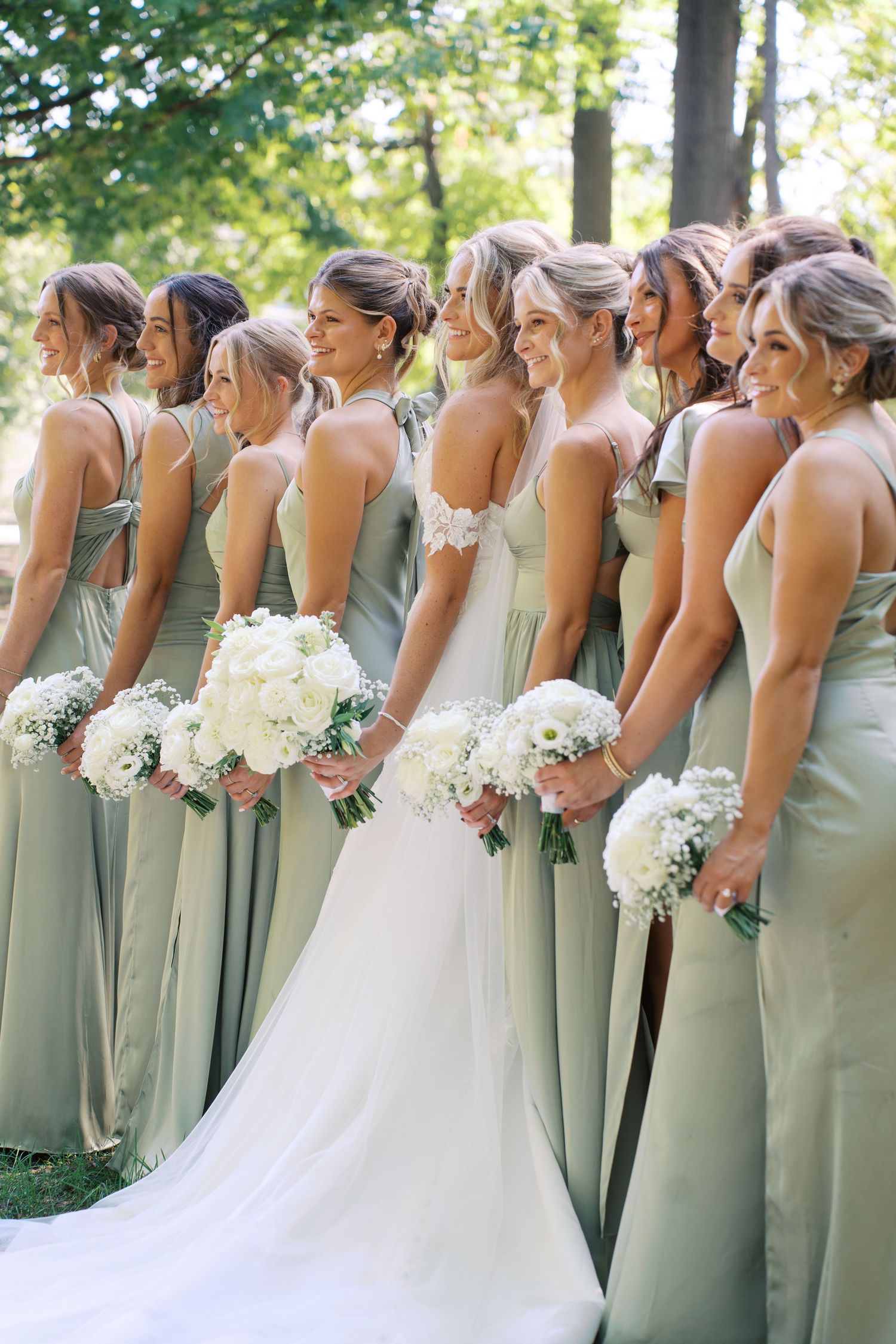 Row of bridesmaids in sage green dresses holding white bouquets pose with bride in outdoor setting.