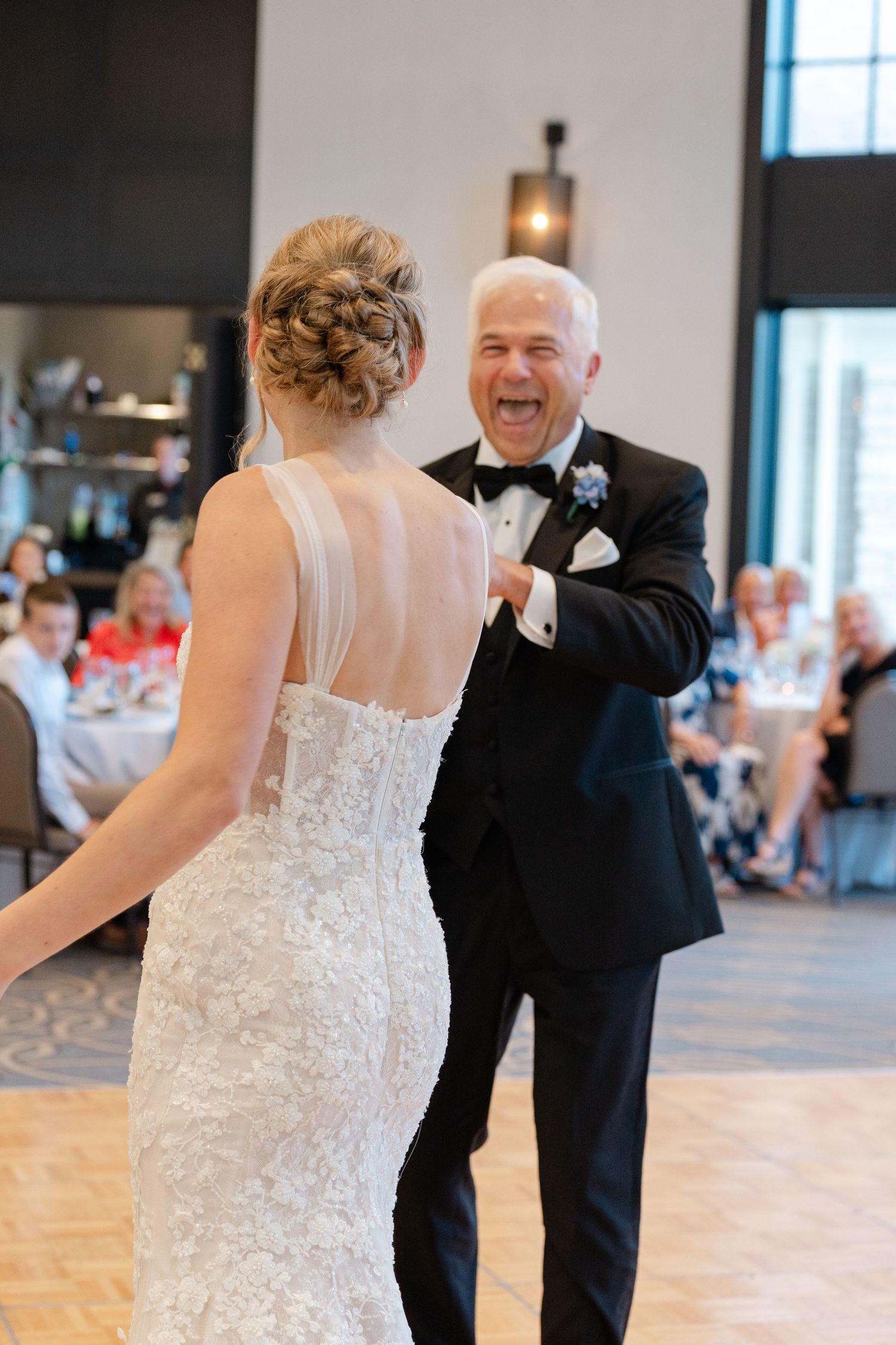 A bride in a lace dress shares a joyful dance with a partner in a black tuxedo at a wedding reception.