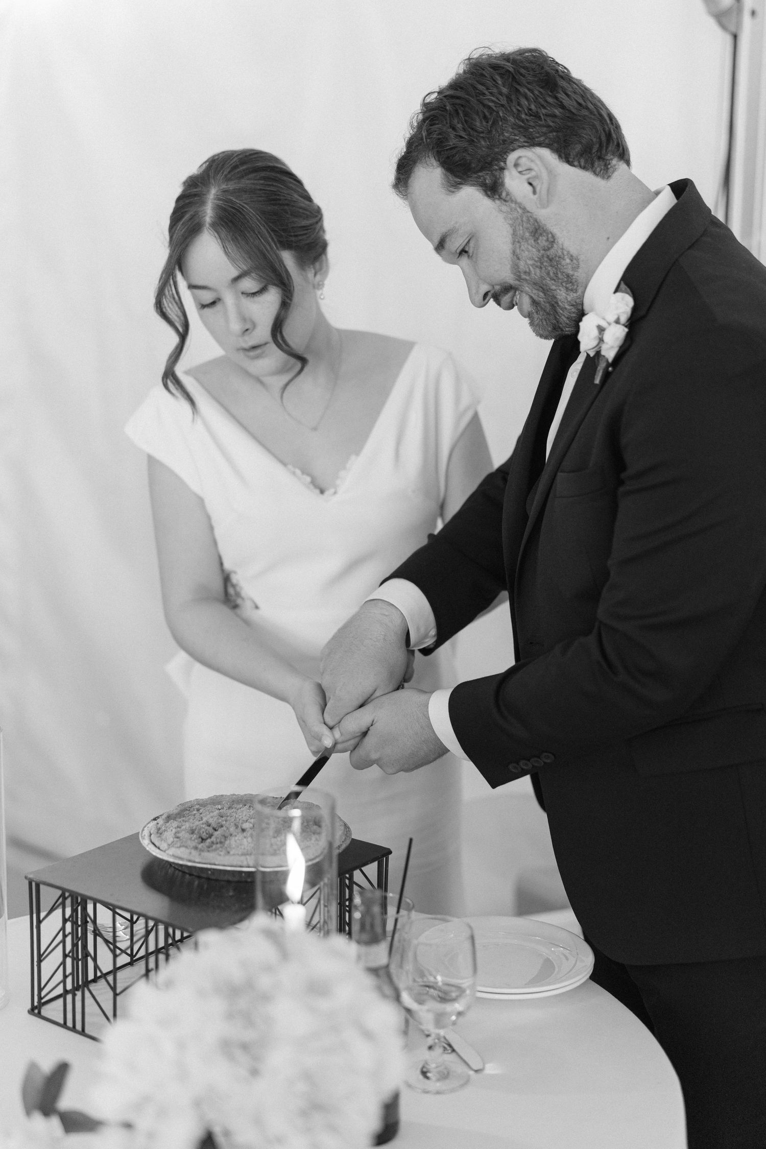 Black and white photo of bride and groom cutting wedding cake together at elegant reception table.