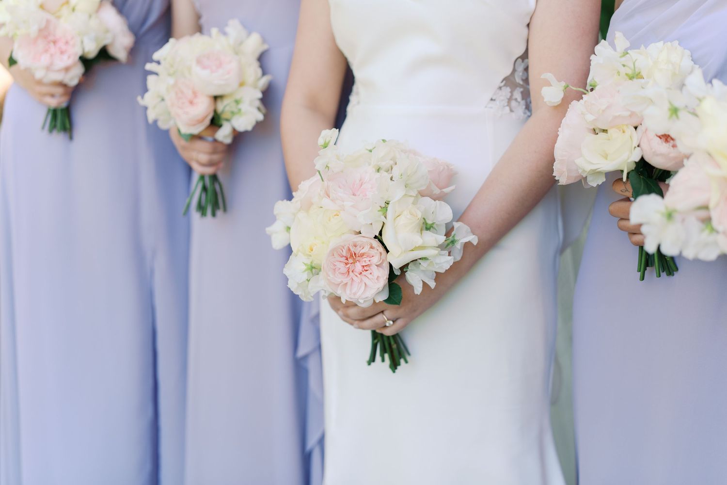 Bride in white gown and bridesmaids in lavender dresses holding soft pink and white floral bouquets.