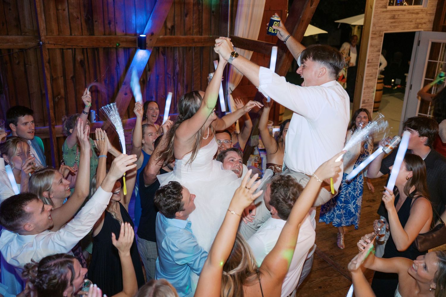Wedding reception party guests dancing with raised arms under blue lighting in a rustic barn venue.