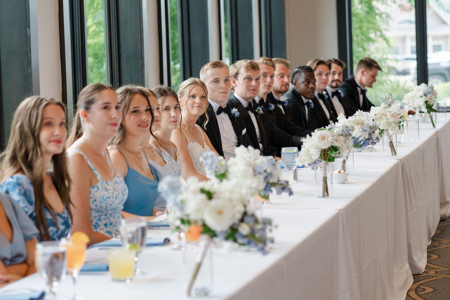 Wedding party seated at long white-clothed table with blue accents and floral centerpieces.