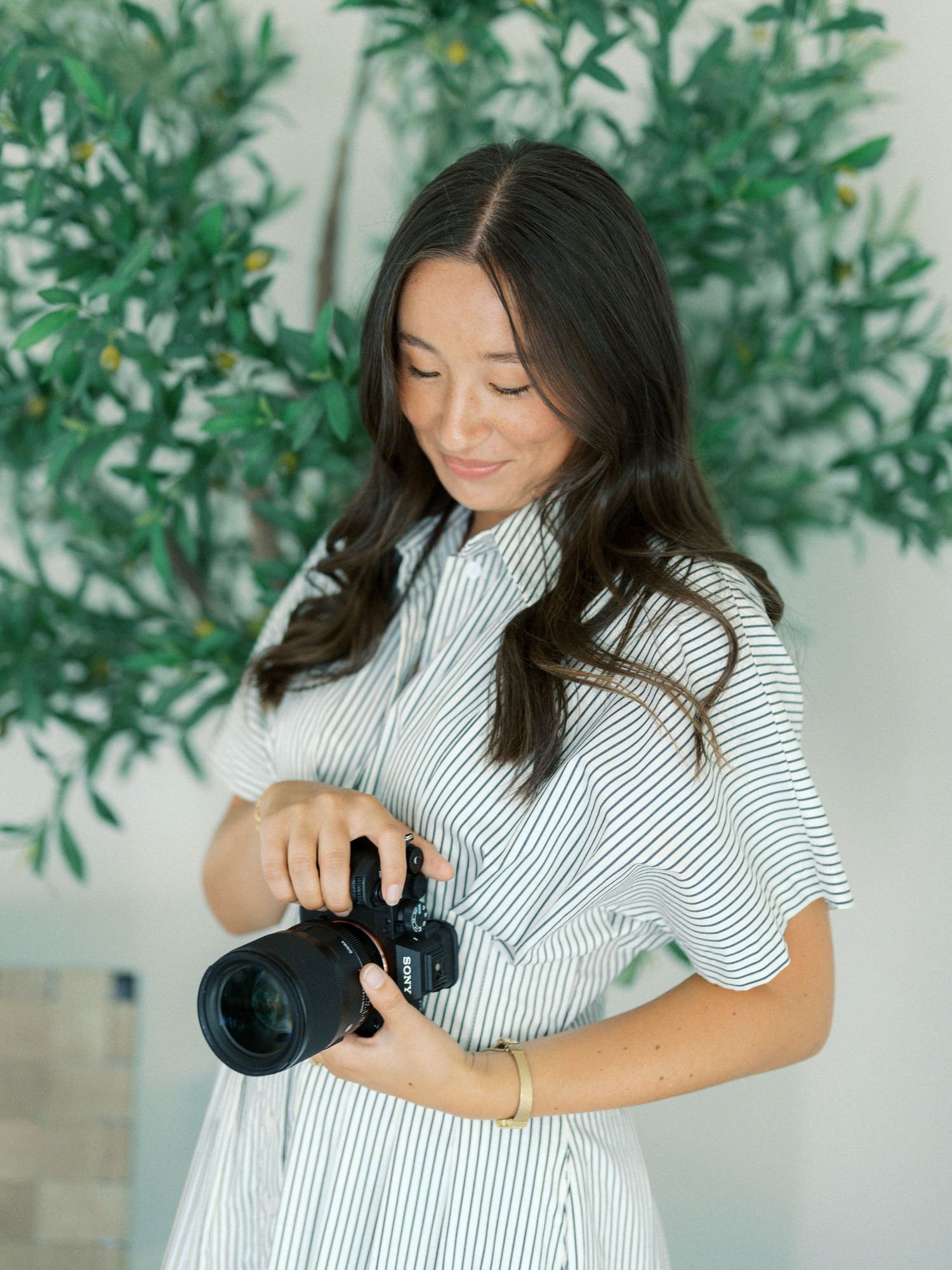Woman in striped dress holding a DSLR camera, smiling, with green olive trees in the background.