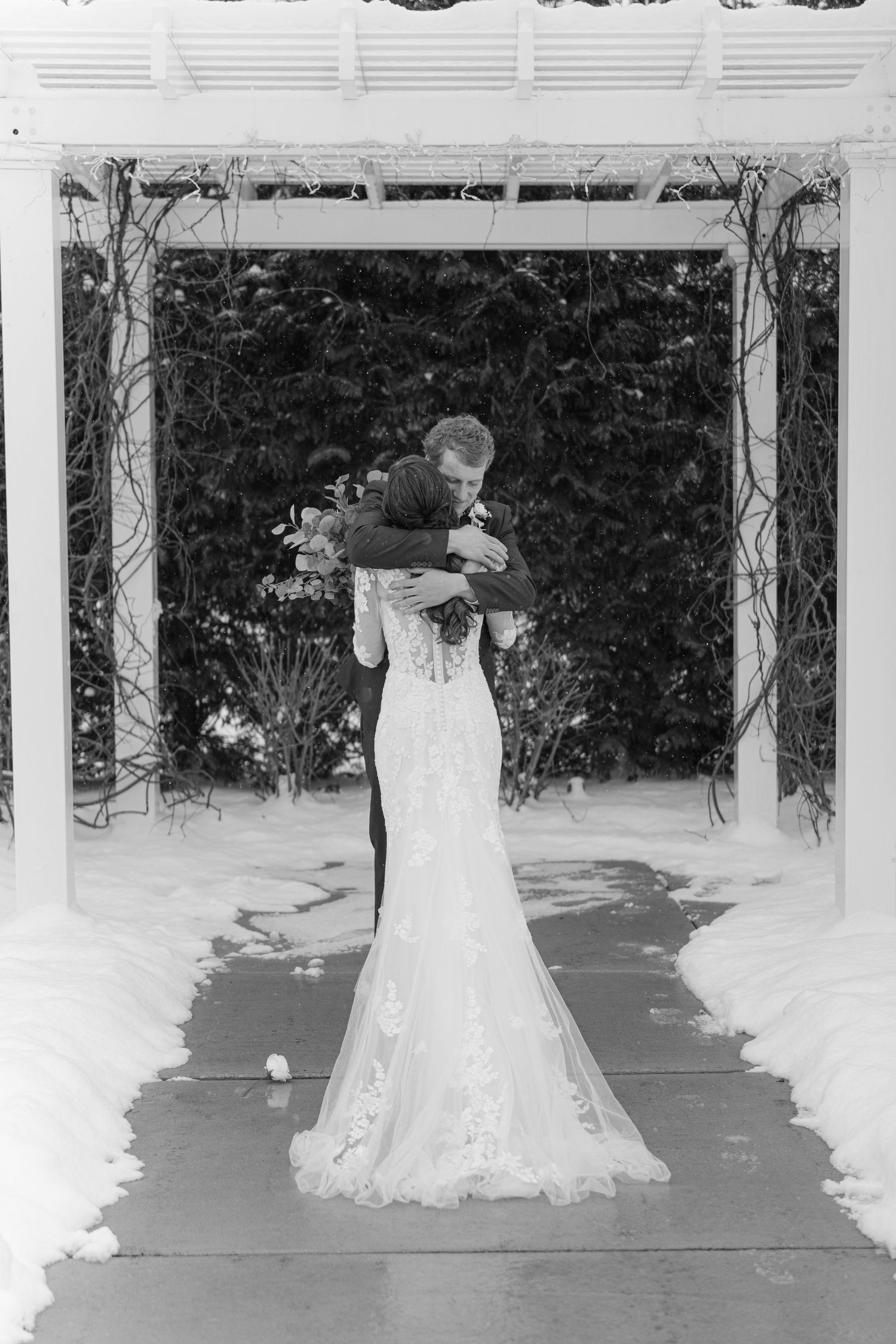 A black and white wedding photo of a couple embracing under a decorated outdoor arbor in winter.
