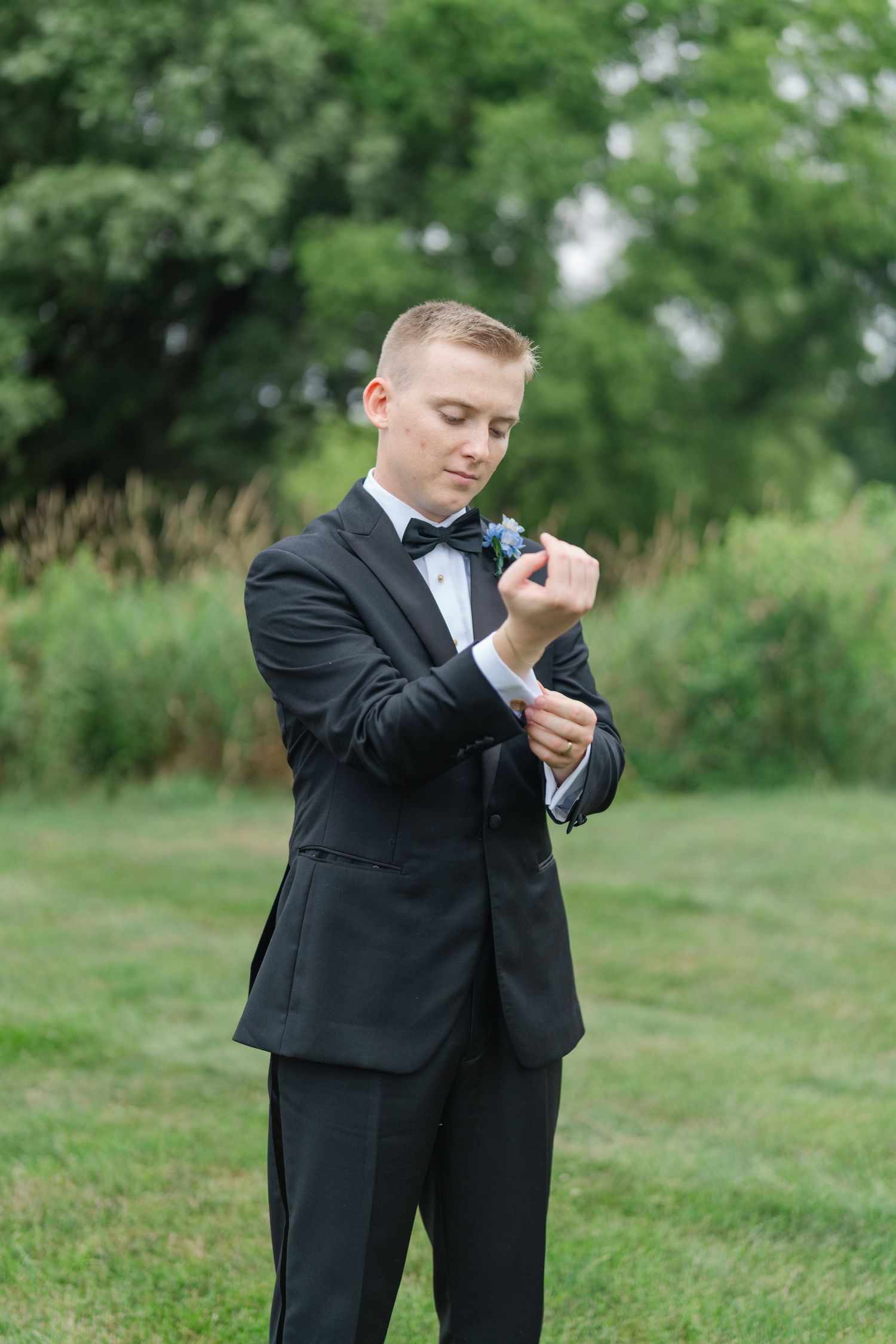 Groom in classic tuxedo strikes playful poses on grassy field during wedding photoshoot.