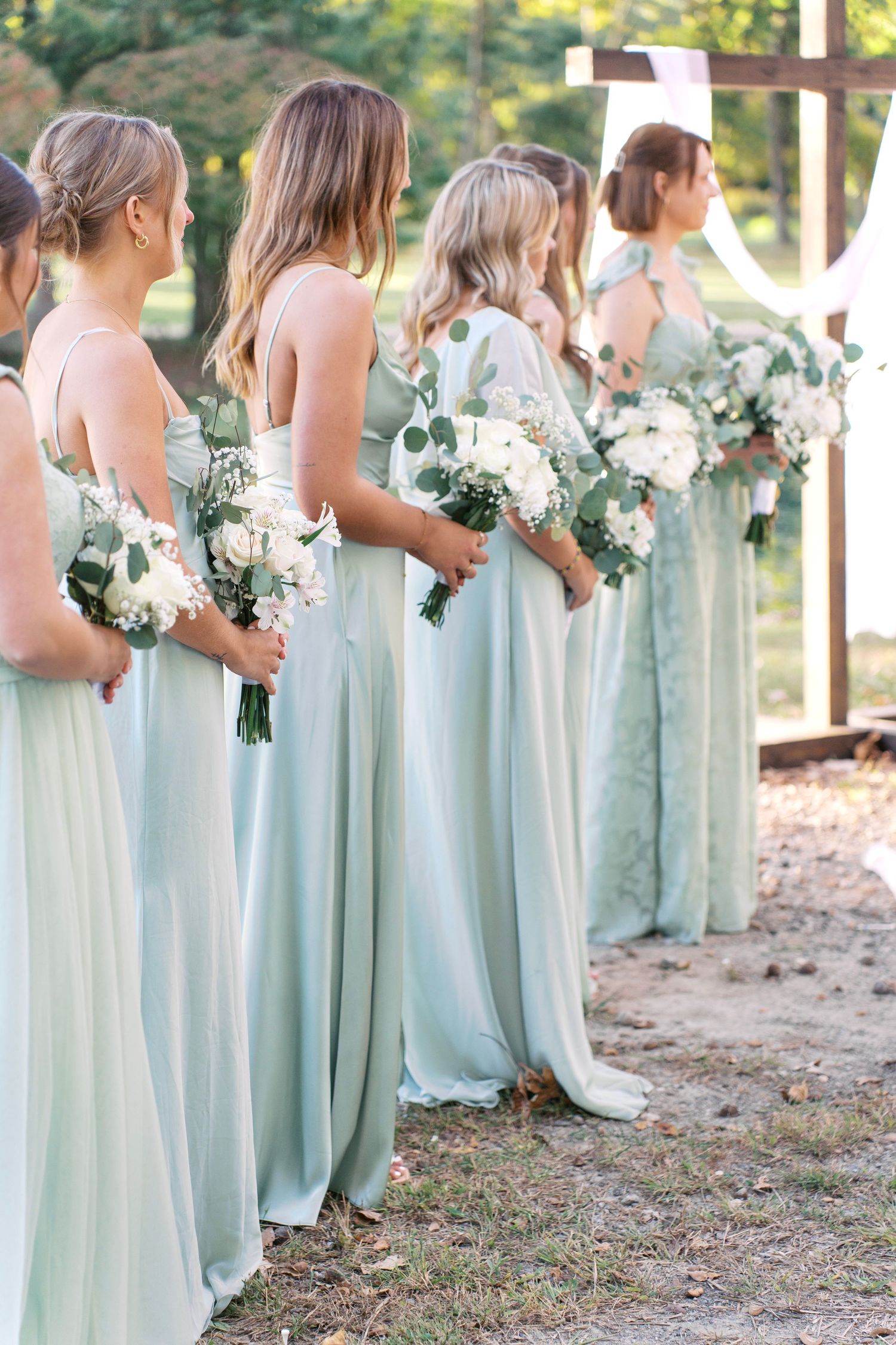 Row of bridesmaids in soft mint dresses holding white bouquets during outdoor ceremony.