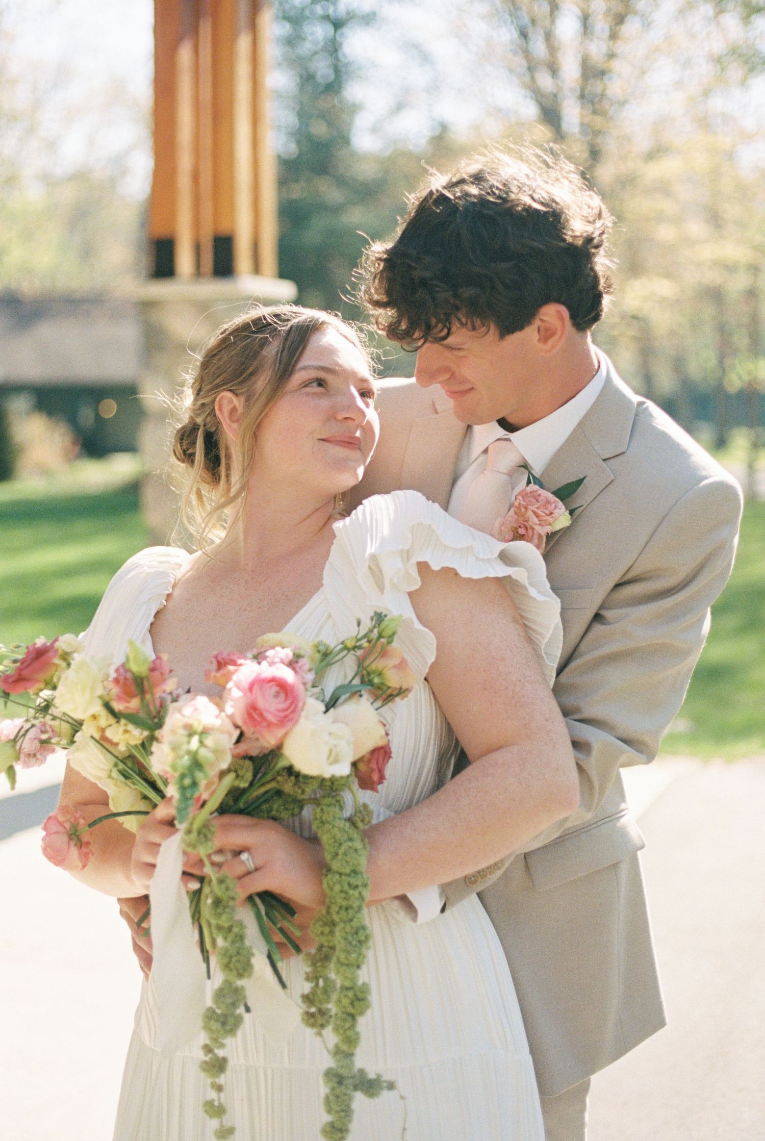 A bride holding a pink floral bouquet embraces her groom in a tan suit outdoors on their wedding day.