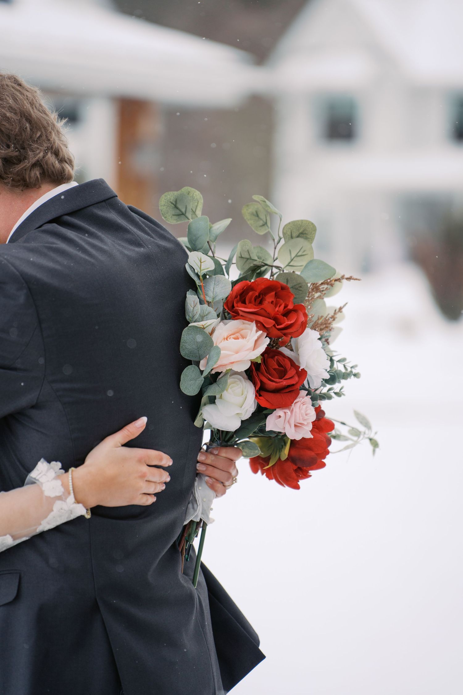 Wedding couple embraces while holding bouquet of red roses and eucalyptus in snow.