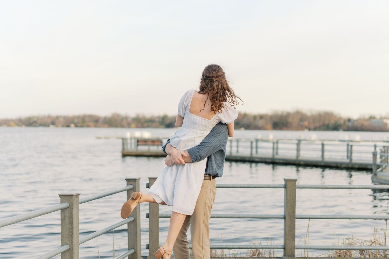 Couple embracing on a lakeside dock at sunset, woman lifted off ground in romantic outdoor engagement photo.