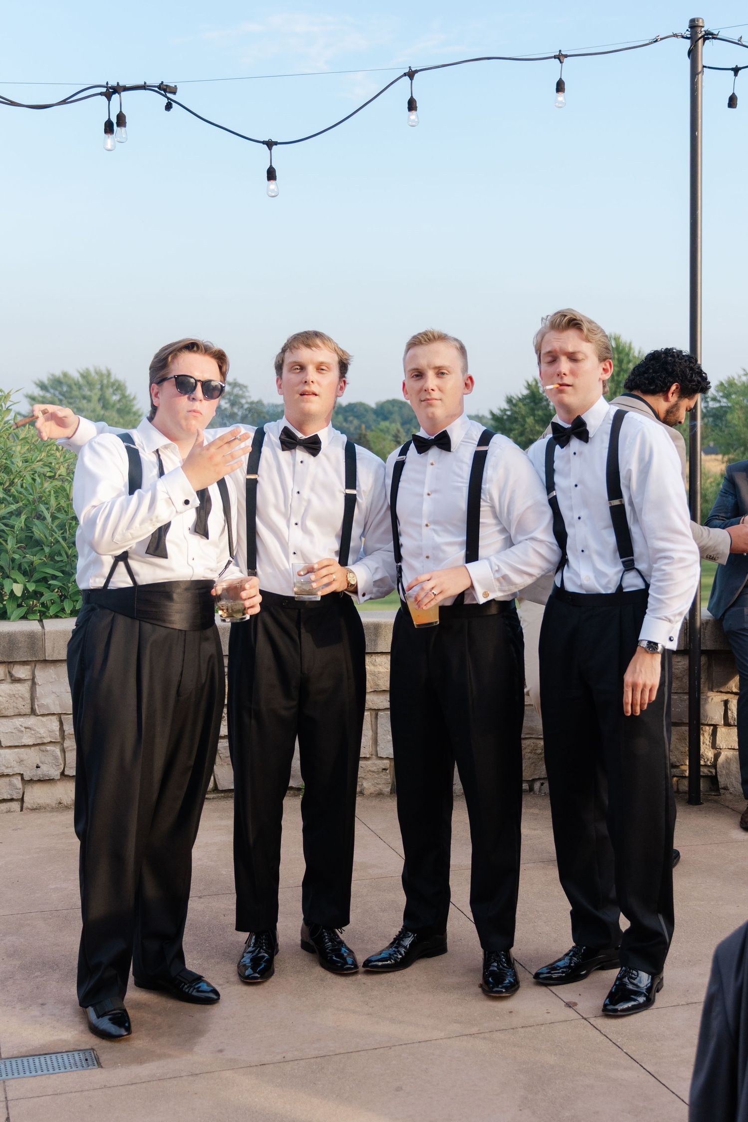 Groomsmen in suspenders and bowties pose together under string lights at an outdoor venue.