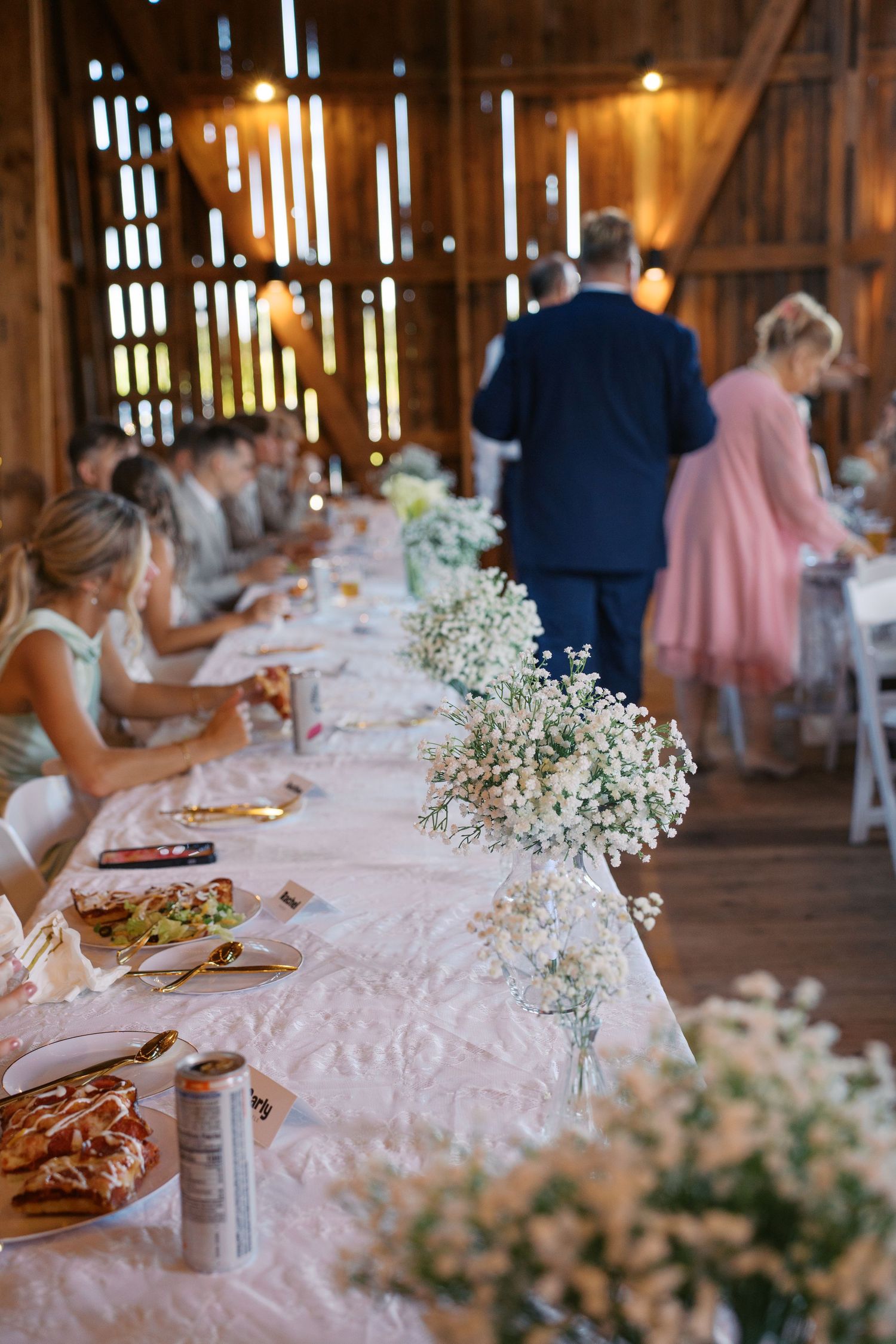 Long reception table decorated with baby's breath flowers and white linens inside a rustic barn venue.